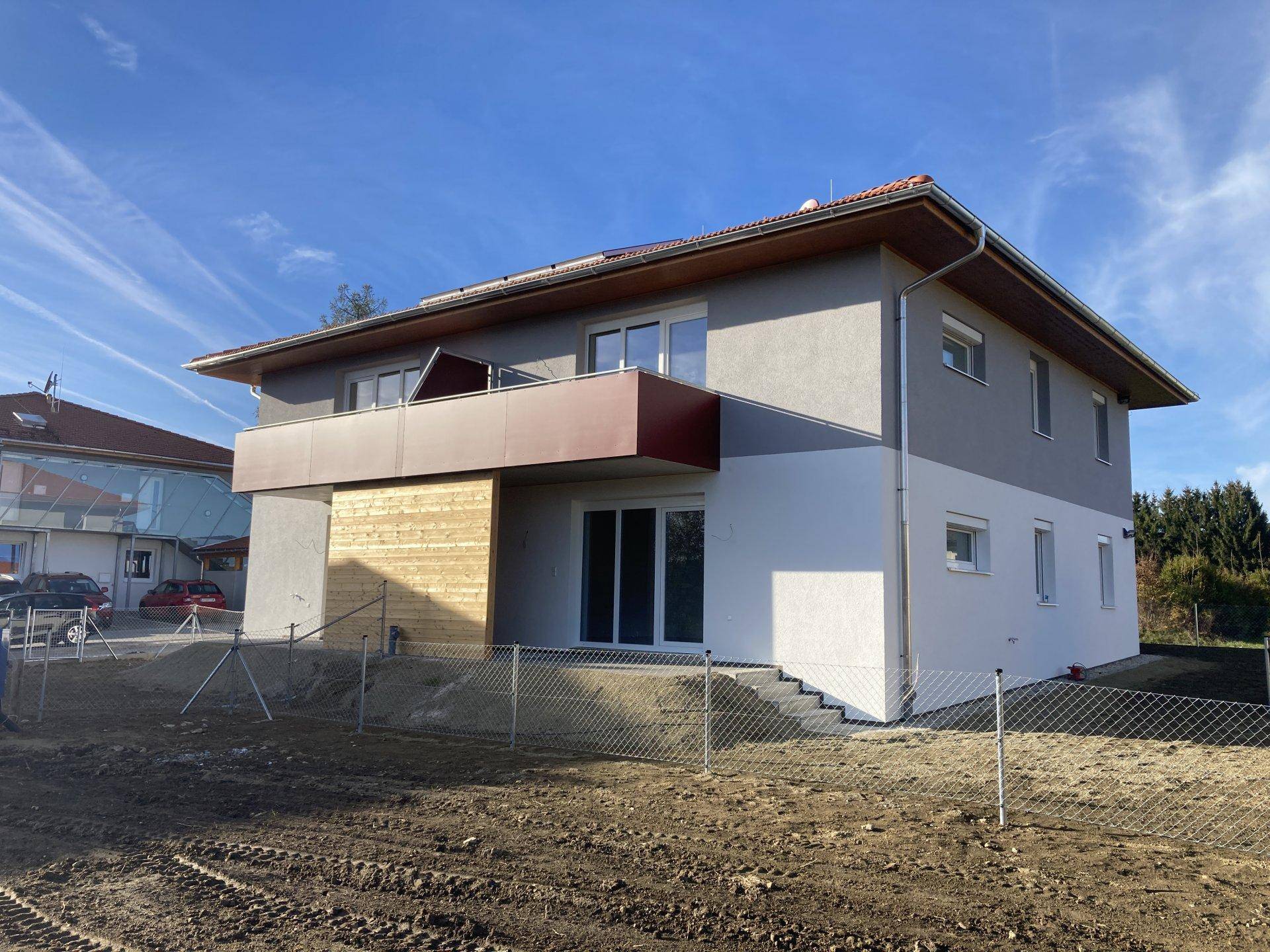 Ansicht des Neubaus von der Seite mit grauer und weißer Fassade, rotem Balkon und umliegendem Baugelände.