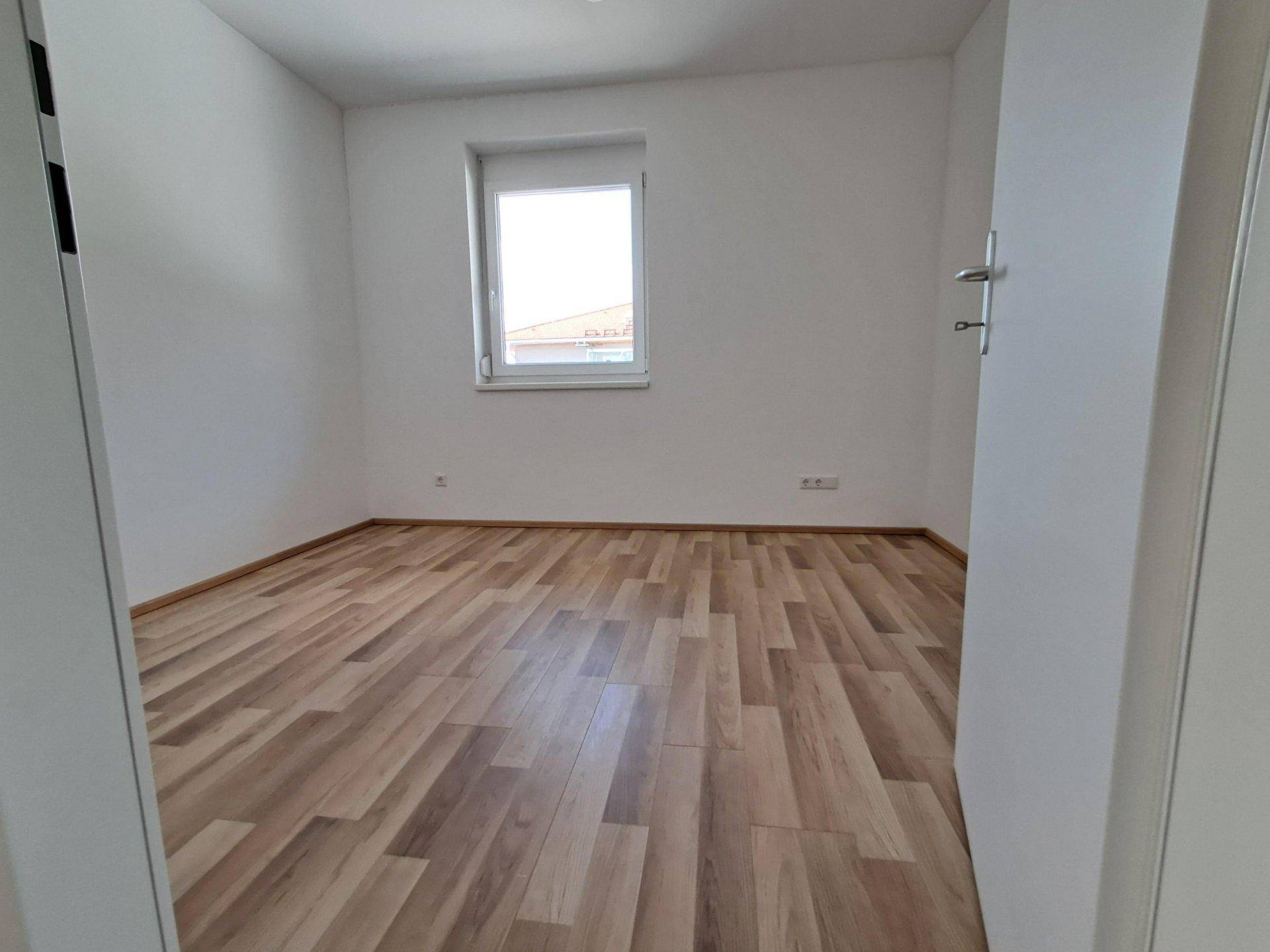 Helles, leeres Schlafzimmer mit Laminatboden, weißen Wänden und einem Fenster.