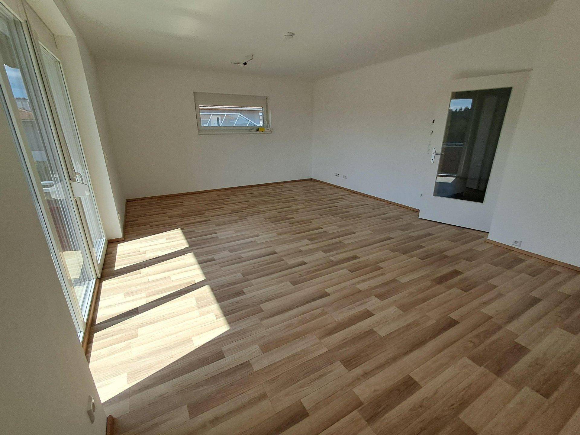 Geräumiger, heller Wohnraum mit Laminatboden und großen Fenstern, die viel Tageslicht hereinlassen.