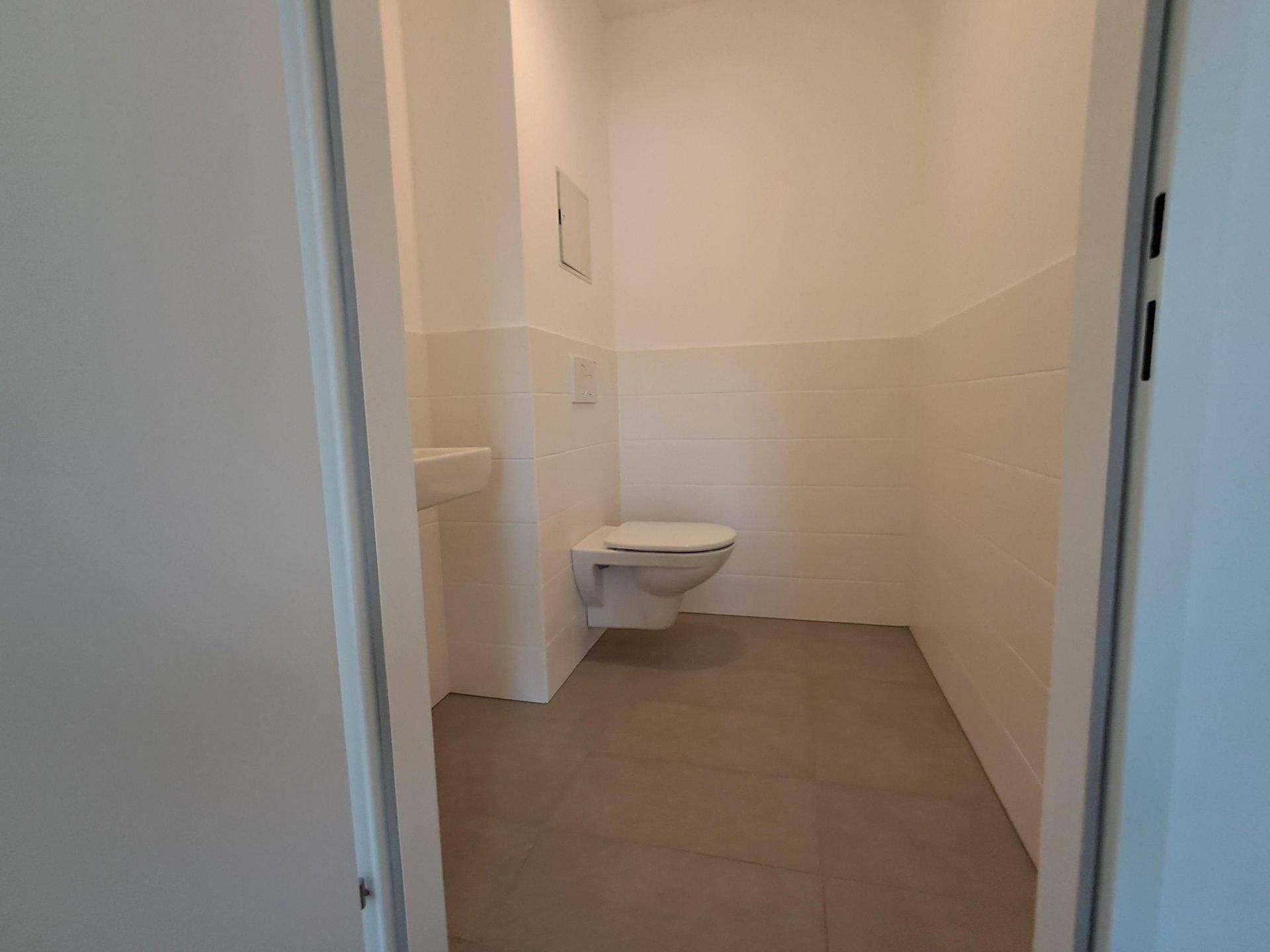 Separates WC mit weißen Fliesen, grauen Bodenfliesen, wandhängendem WC und kleinem Waschbecken.