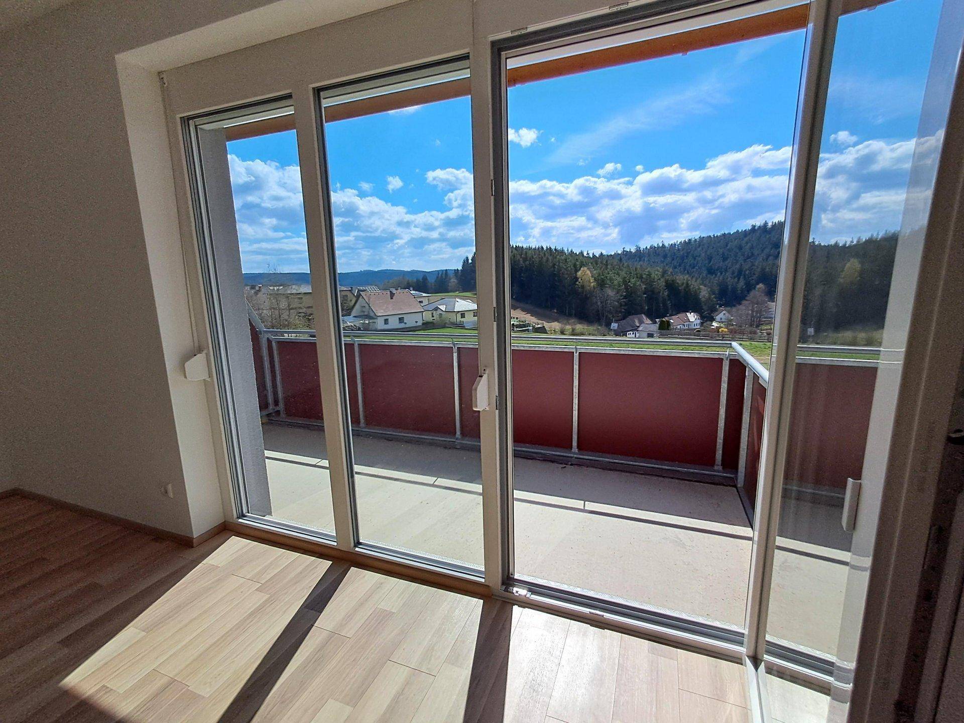 Blick von innen durch große Glasschiebetüren auf einen Balkon mit rotem Geländer und grüne Landschaft.