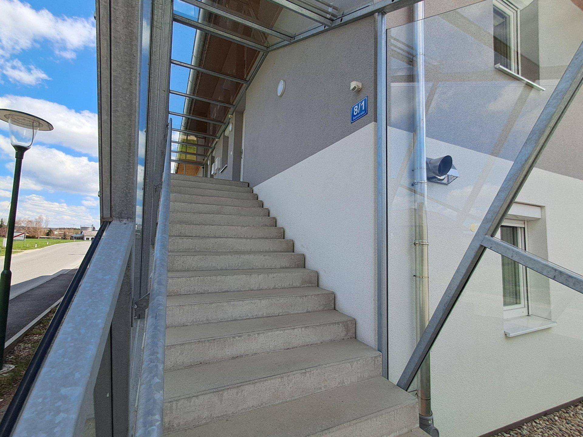 Außentreppe mit Betonstufen und Glasgeländer, die zum Eingang des Gebäudes führt.