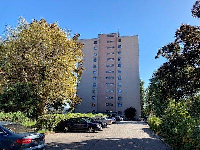 Hohes Wohngebäude mit Parkplätzen und Bäumen im Vordergrund unter klarem Himmel.