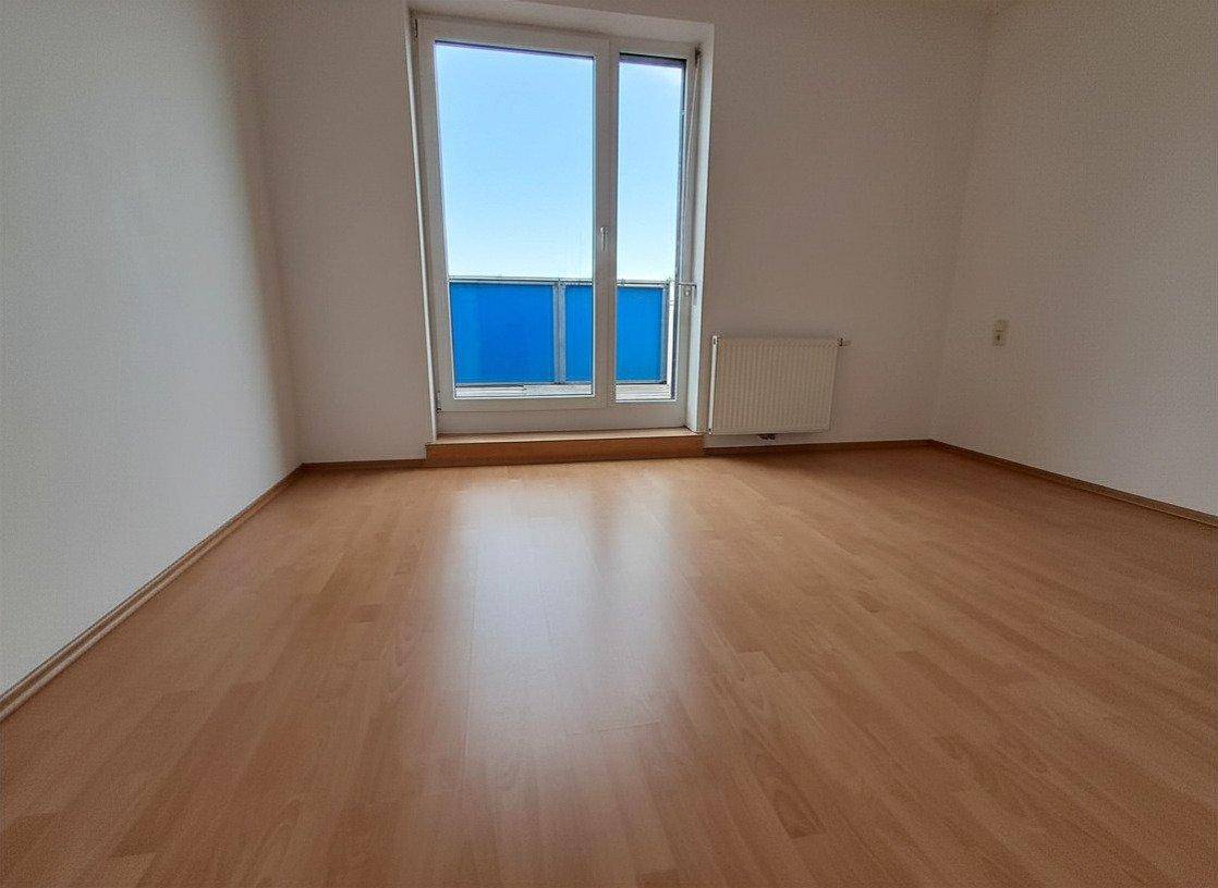 Leeres Zimmer mit Laminatboden und einer großen Fenstertür, die viel Licht spendet.