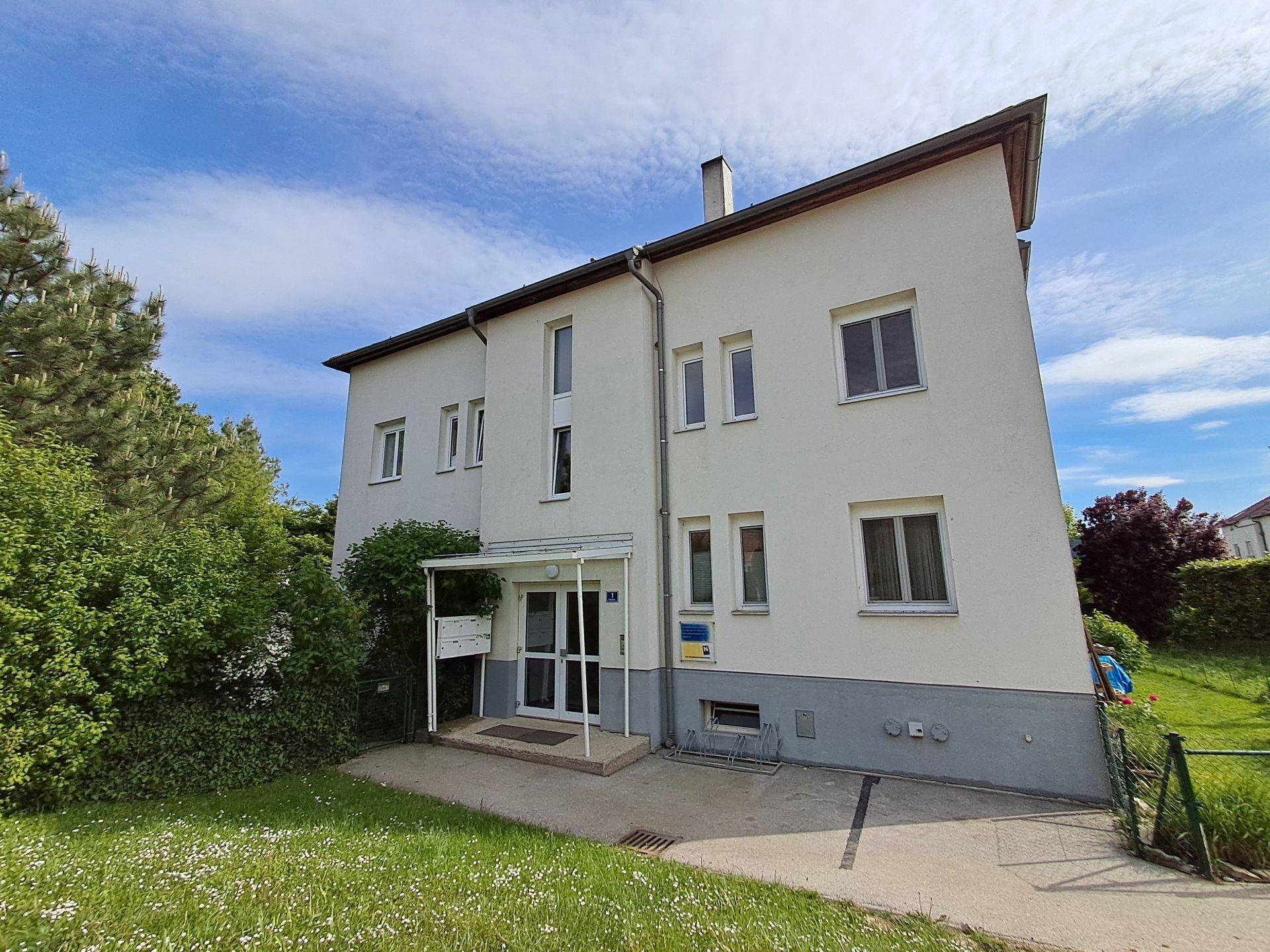 Vorderansicht eines gepflegten Mehrfamilienhauses mit Eingangsbereich und umliegenden Grünflächen bei Sonnenschein.
