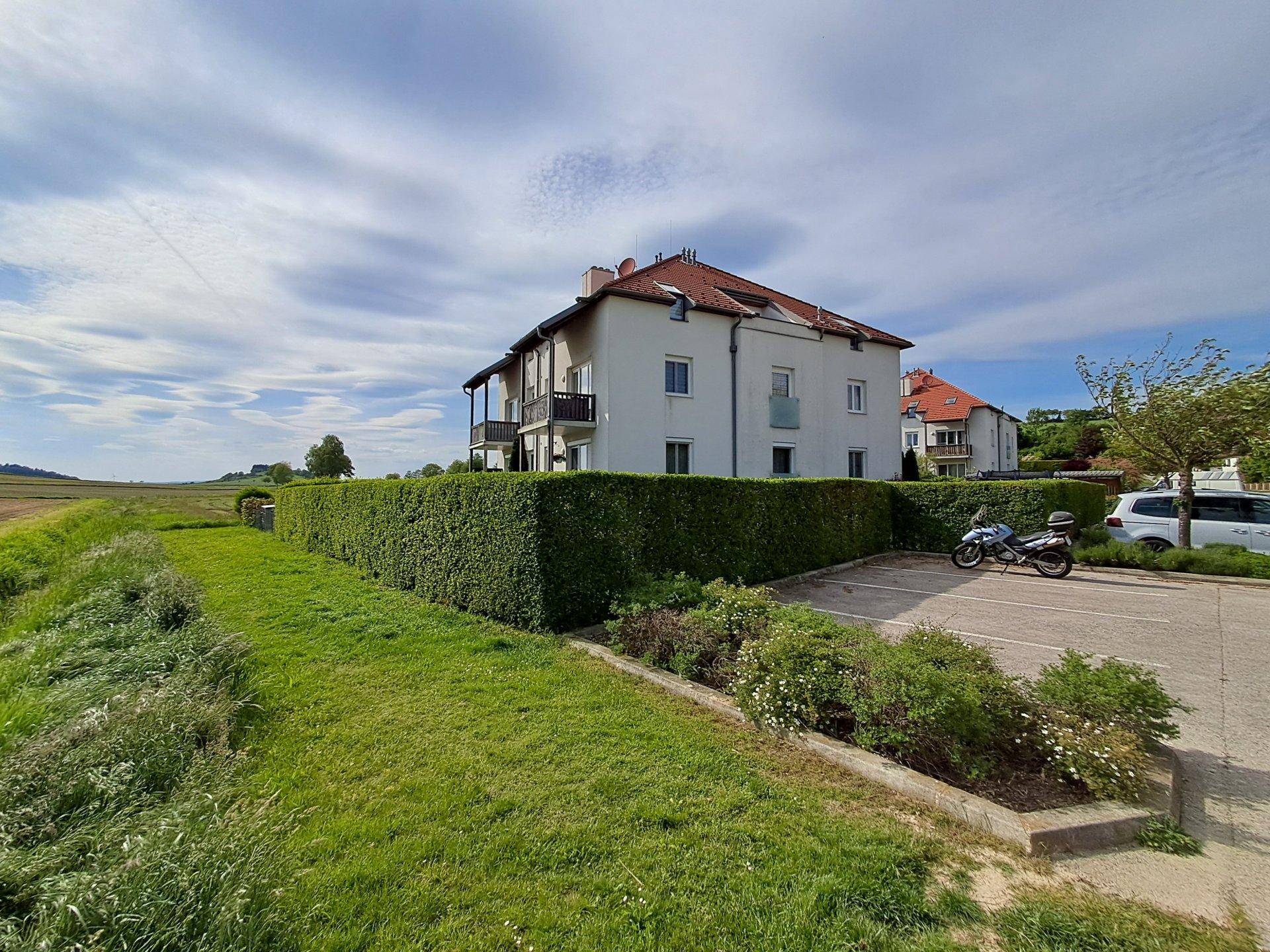 Mehrfamilienhaus mit Parkplätzen, einer hohen Hecke und weitem Blick über die ländliche Landschaft.