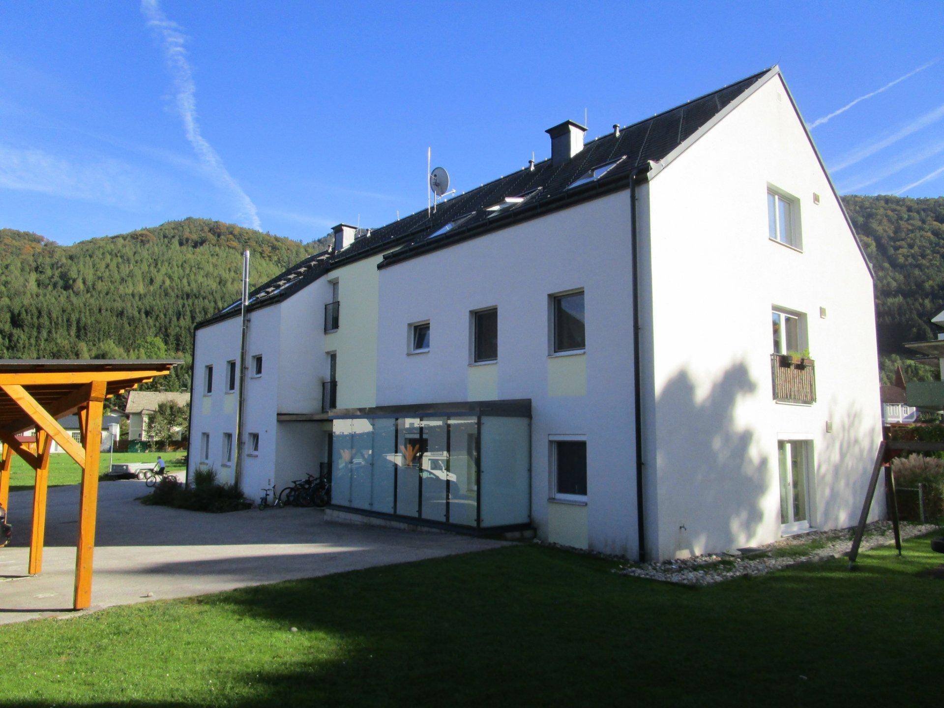 Weiße Fassade eines Mehrfamilienhauses mit Carport und Grünfläche vor bewaldeten Hügeln.