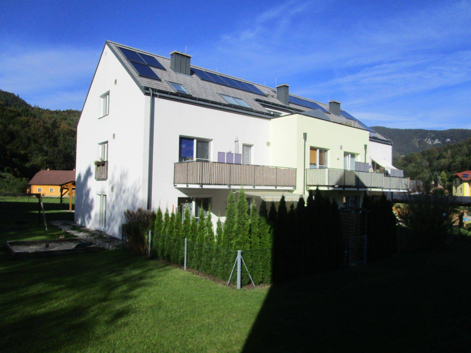 Rückseite eines modernen Mehrfamilienhauses mit Solaranlage und gepflegtem Rasenbereich in der Sonne.
