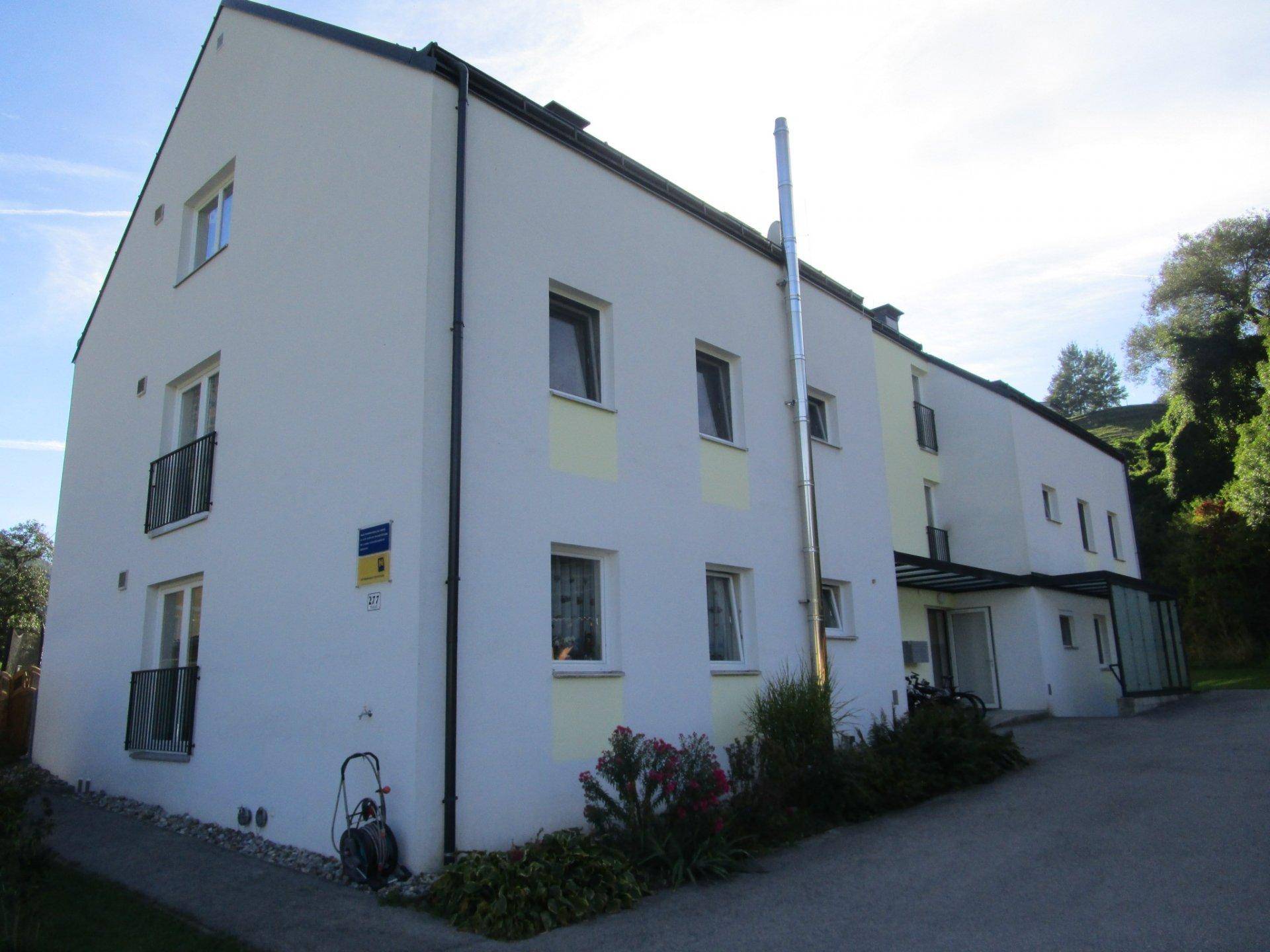 Seitenansicht eines weißen Mehrfamilienhauses mit Balkonen und einem langen Kaminrohr an der Fassade.