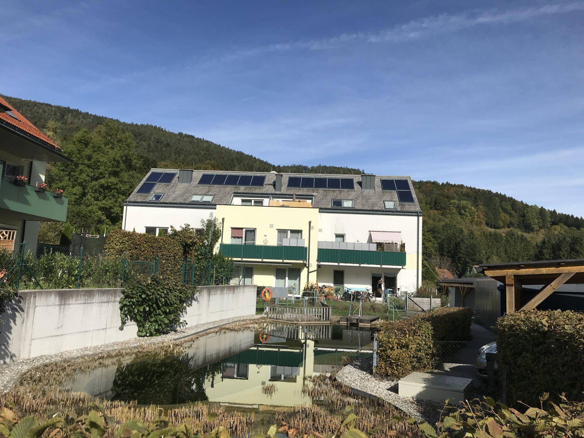 Modernes Mehrfamilienhaus mit Solaranlage auf dem Dach und einem Teich im Gartenbereich unter blauem Himmel.