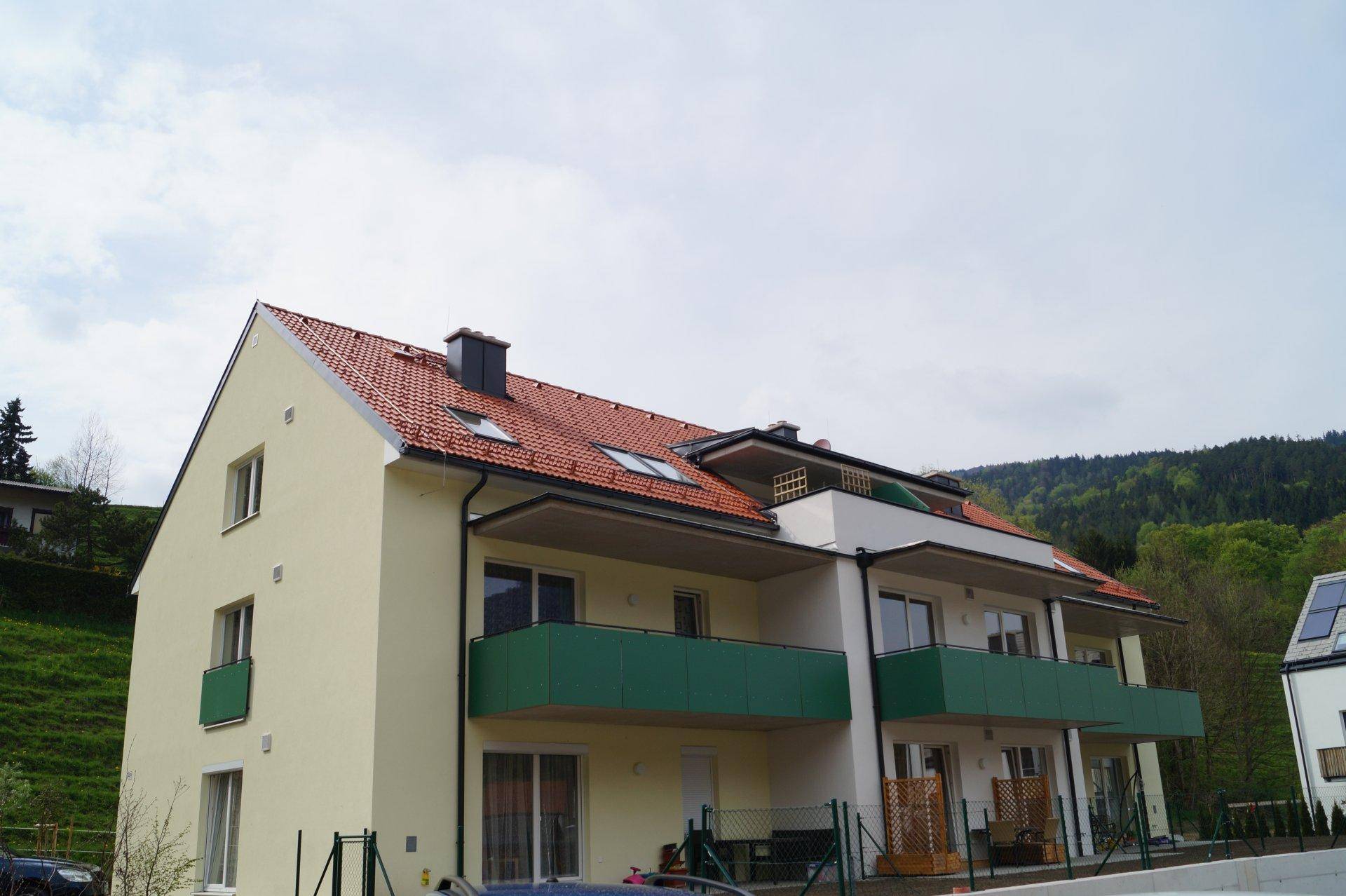 Modernes Mehrfamilienhaus mit rotem Ziegeldach, grünen Balkonen und heller Fassade in ländlicher Umgebung.