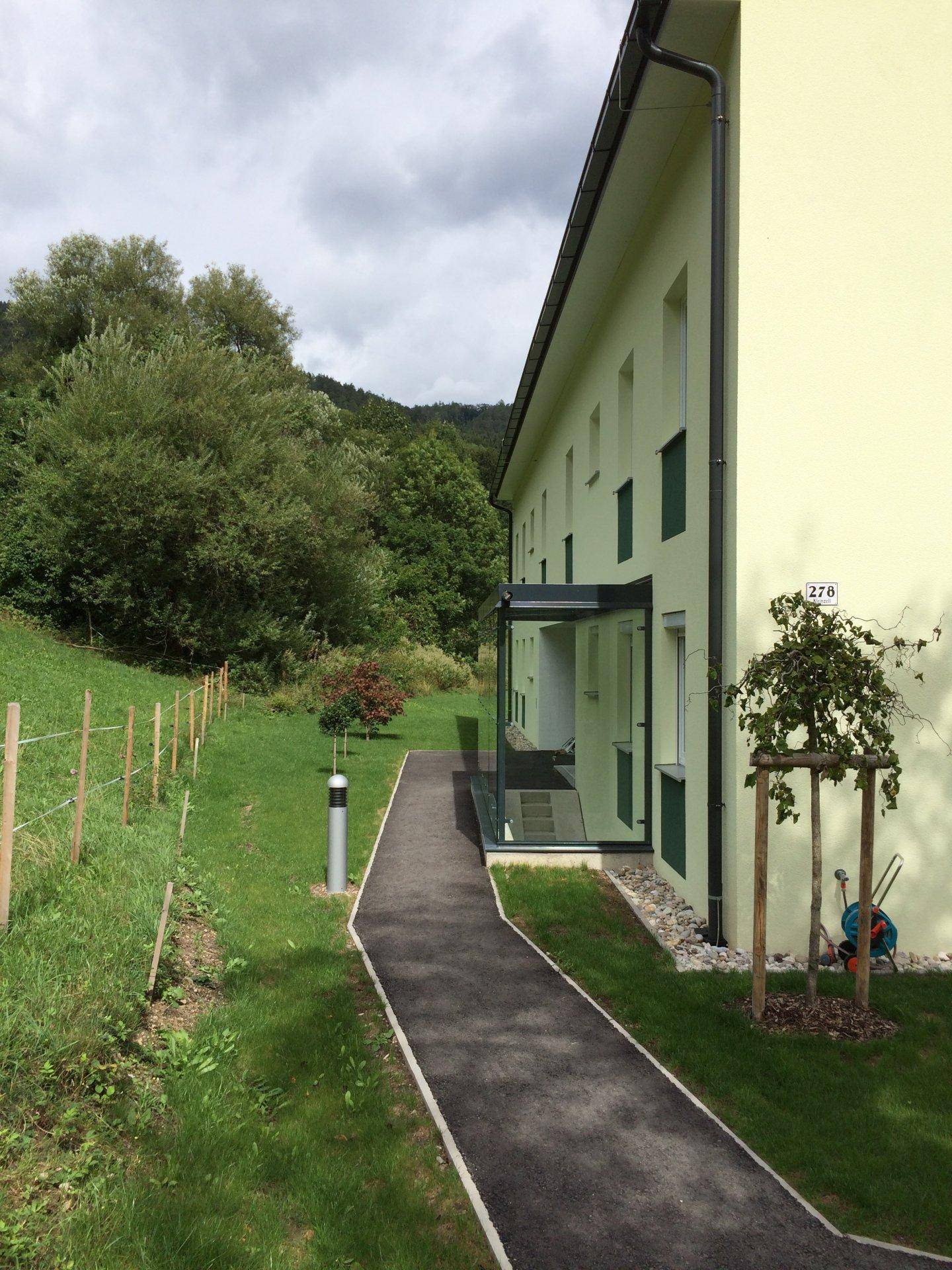Seitenansicht des Gebäudes mit einem Gehweg, umgeben von grünen Wiesen und Bäumen.