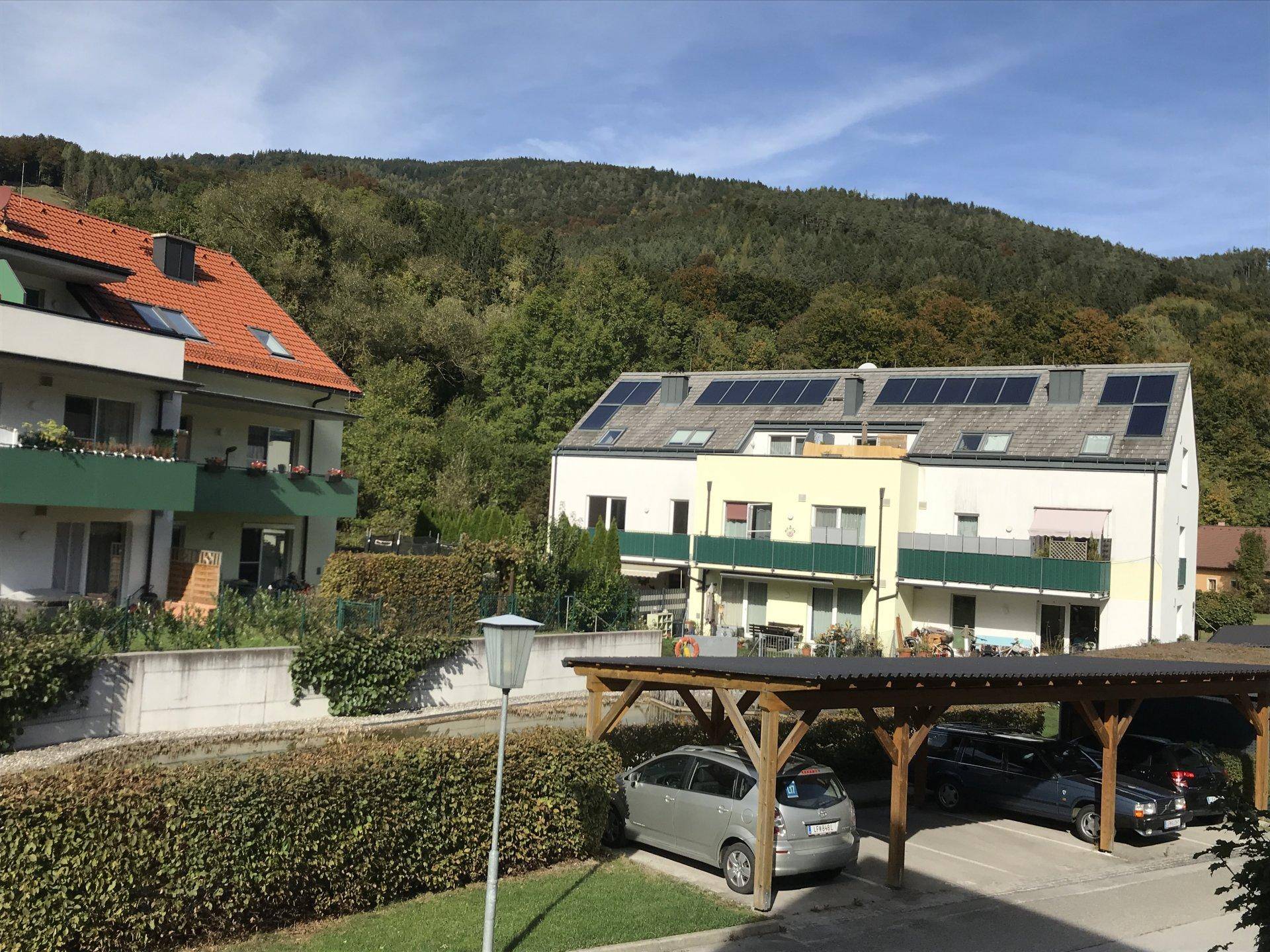 Zwei Mehrfamilienhäuser mit Solaranlagen und einem Carport vor bewaldeten Hügeln an einem sonnigen Tag.