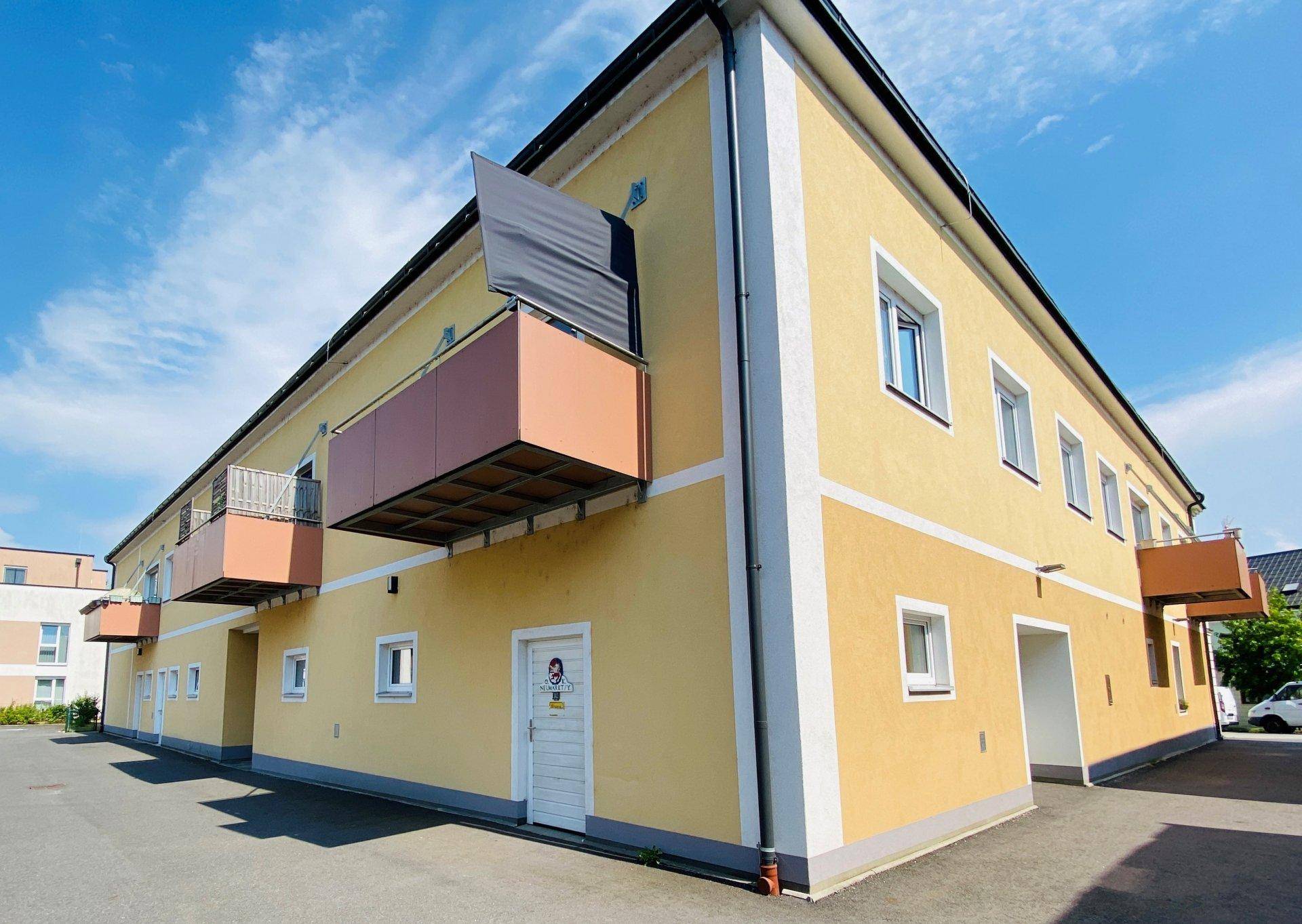 Gelbe Fassade eines Wohngebäudes mit mehreren Balkonen und Parkplätzen davor unter blauem Himmel.