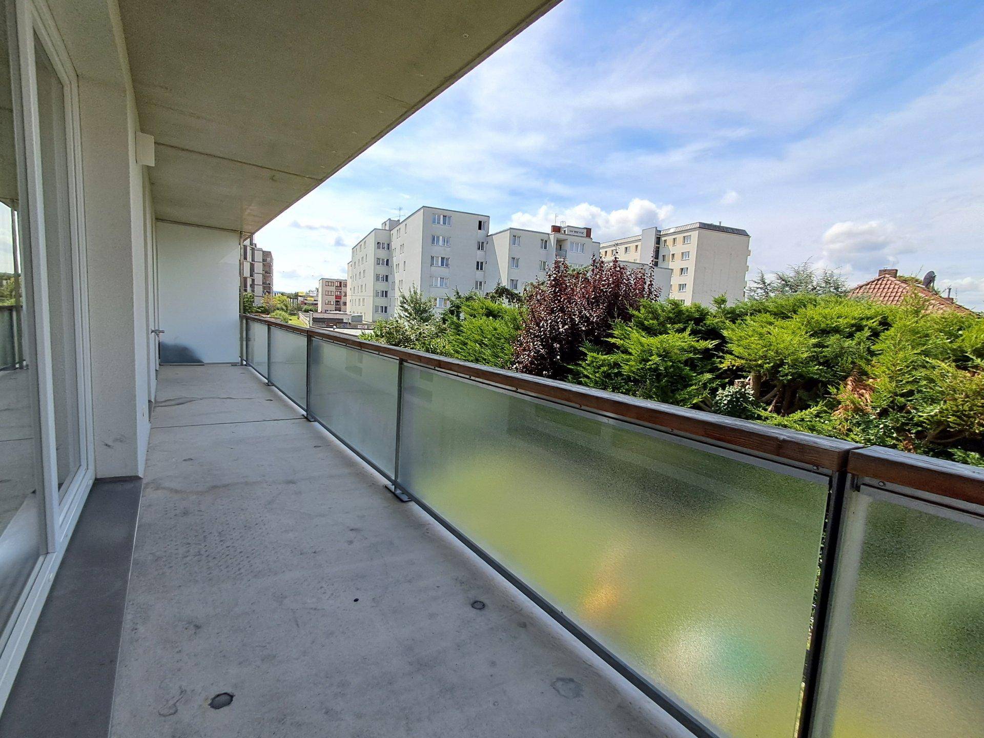 Langer Balkon mit Milchglasgeländer und Blick auf Bäume sowie weitere Wohngebäude.