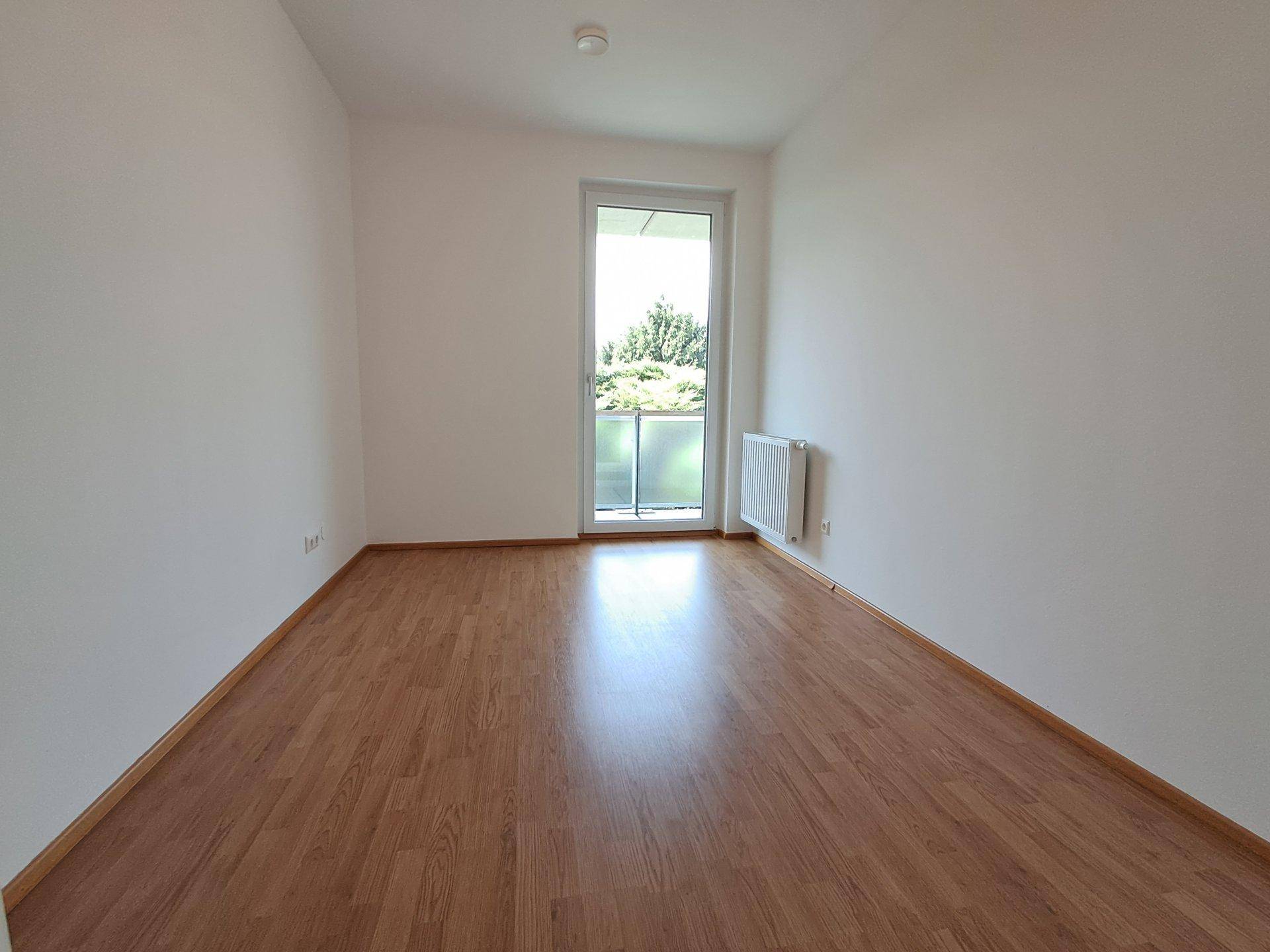 Leeres Zimmer mit Laminatboden, weißen Wänden und Balkonzugang, ausgestattet mit einem Heizkörper.