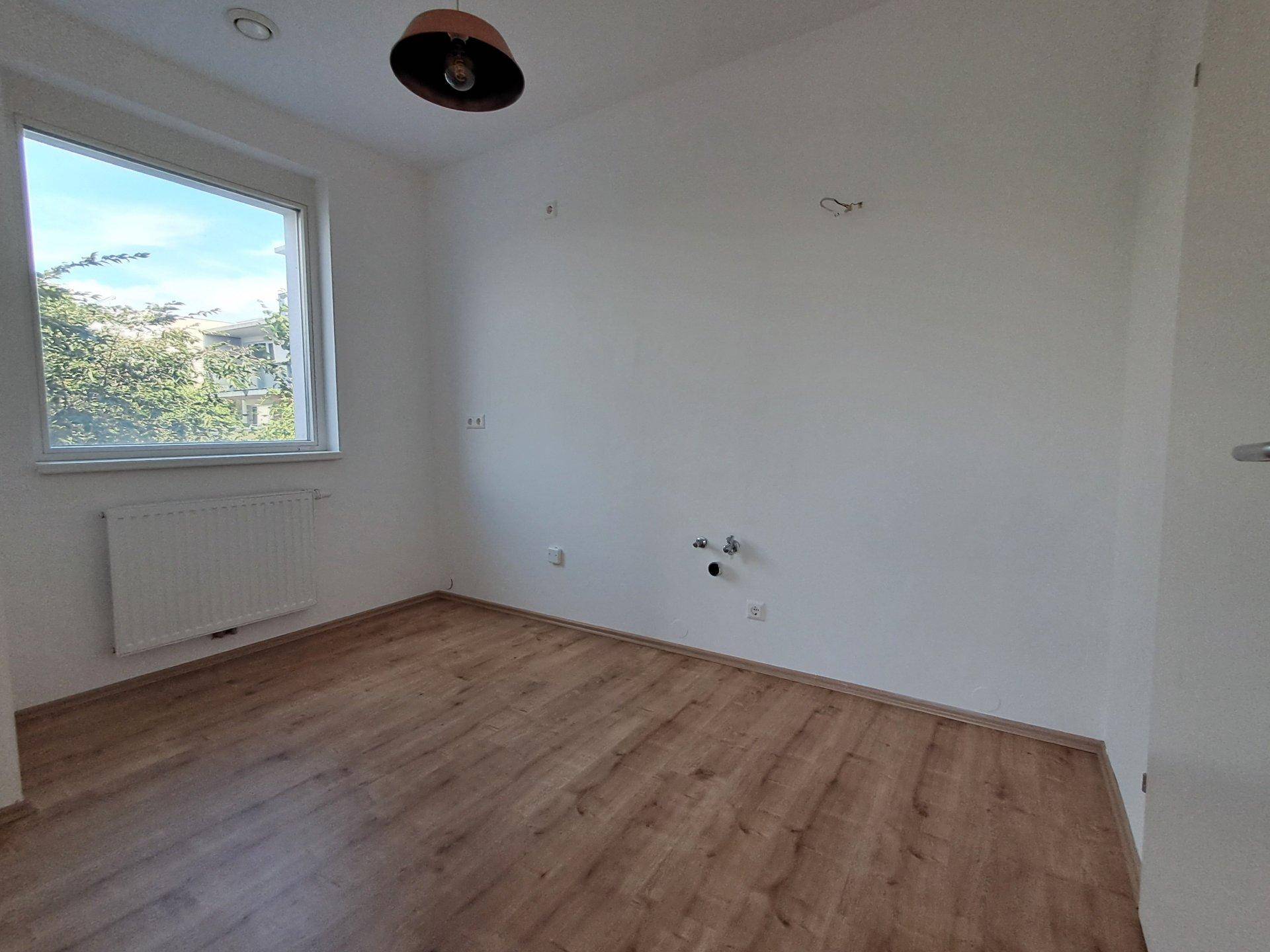 Leerer Raum mit Laminatboden, weiß gestrichenen Wänden und einem Fenster, mit sichtbaren Anschlüssen.