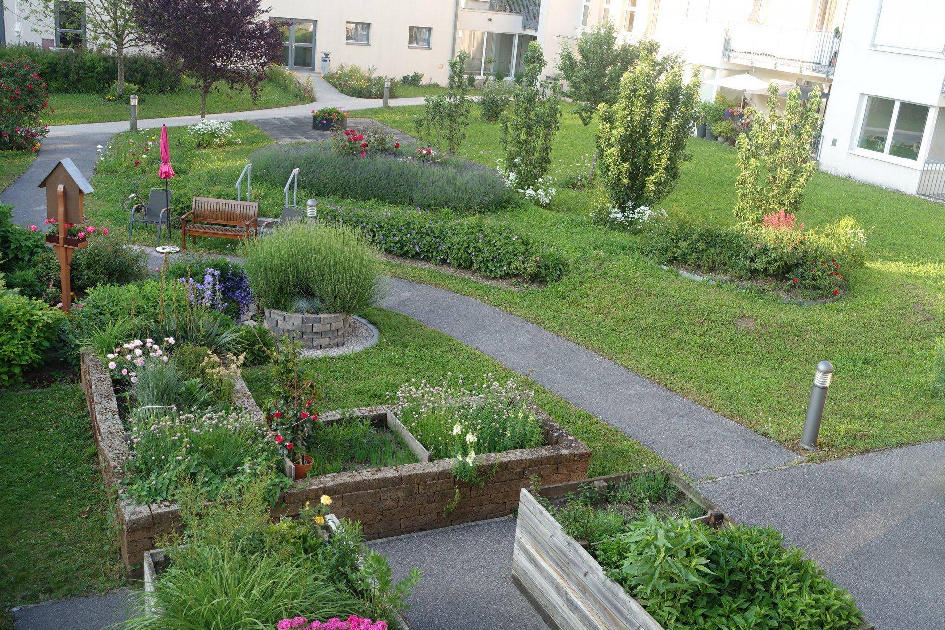 Gepflegter Gemeinschaftsgarten mit Hochbeeten, Blumen, Sträuchern und Sitzgelegenheiten.