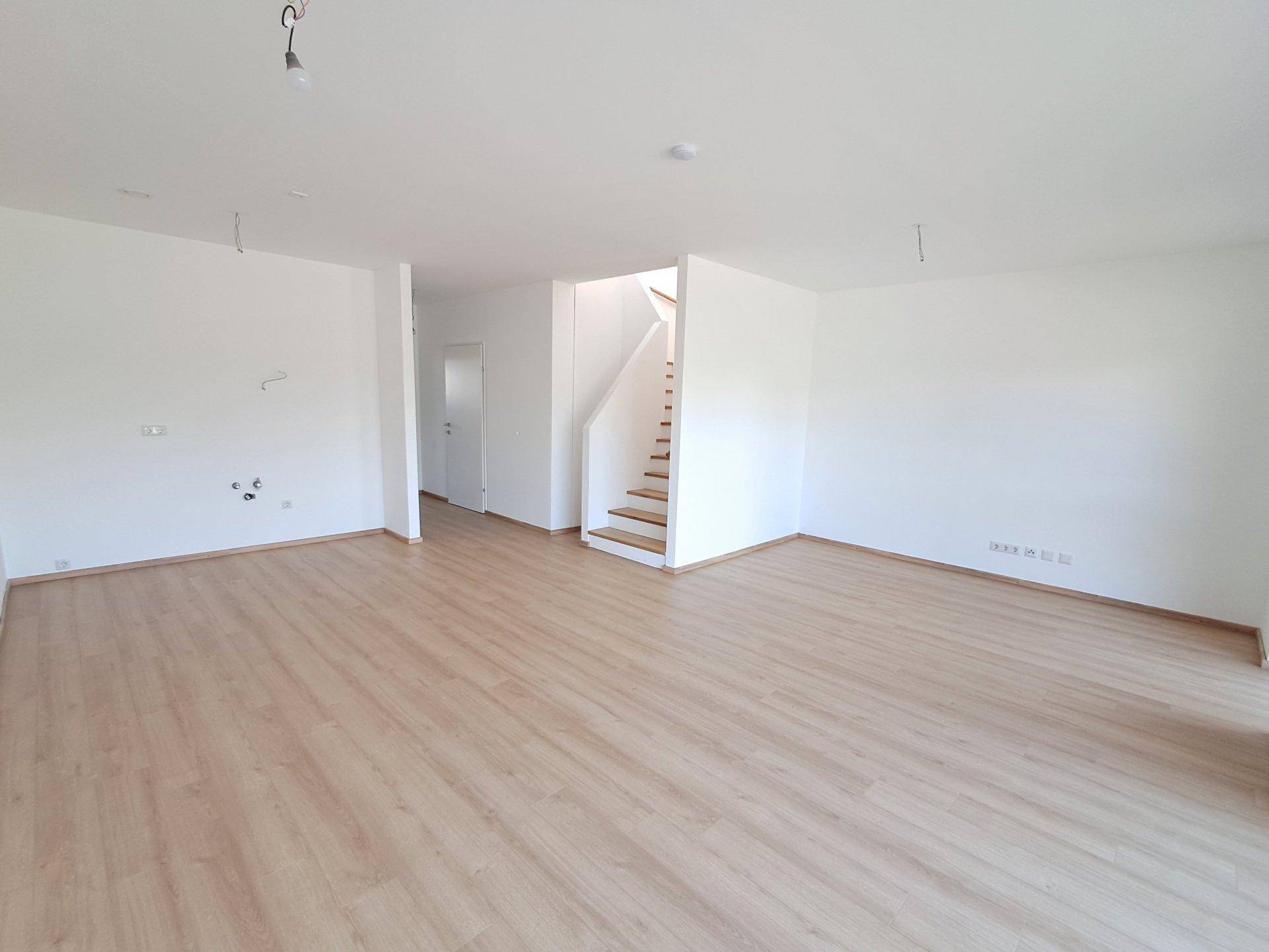 Geräumiger, leerer Wohnbereich mit Laminatboden und offener Treppe zum Obergeschoss.