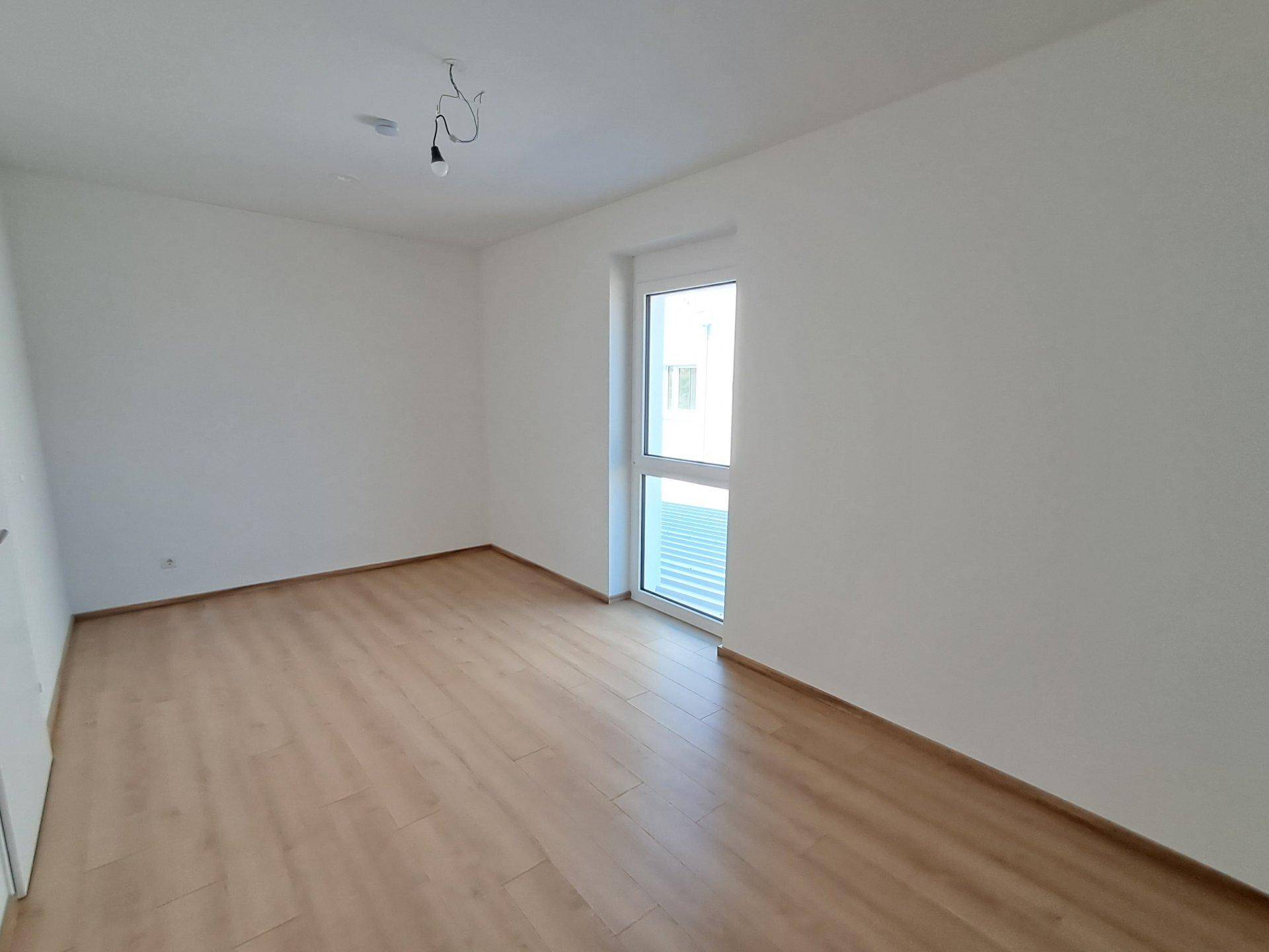Leeres Schlafzimmer mit Laminatboden und einem Fenster, das viel Licht hereinlässt.
