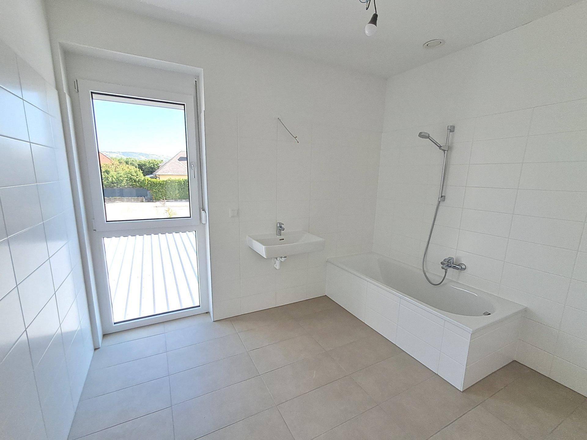 Helles Badezimmer mit Badewanne, Waschbecken, Fliesenboden und Fenster.