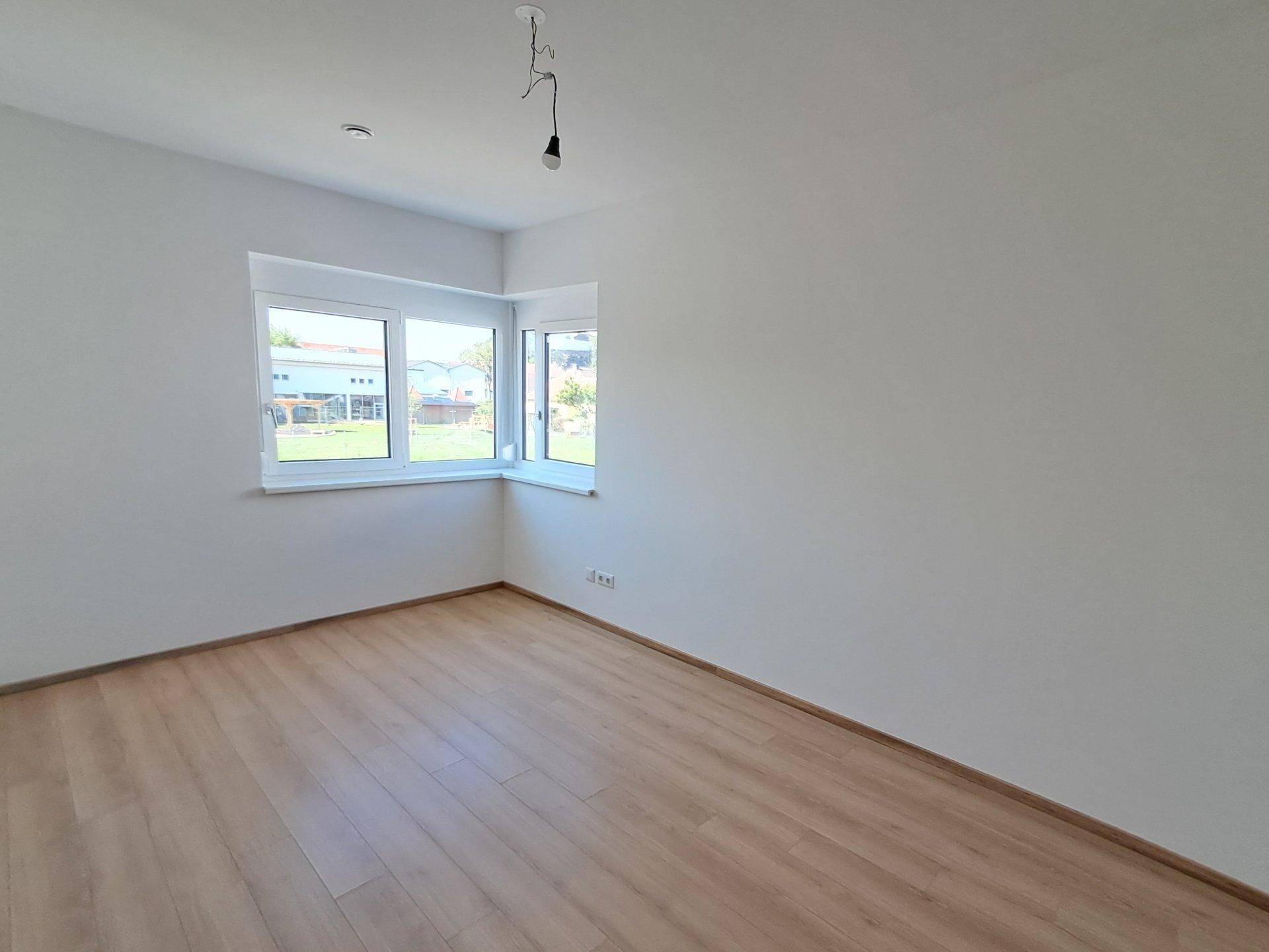Helles, leeres Zimmer mit Laminatboden und Eckfenstern, ideal als Kinderzimmer.