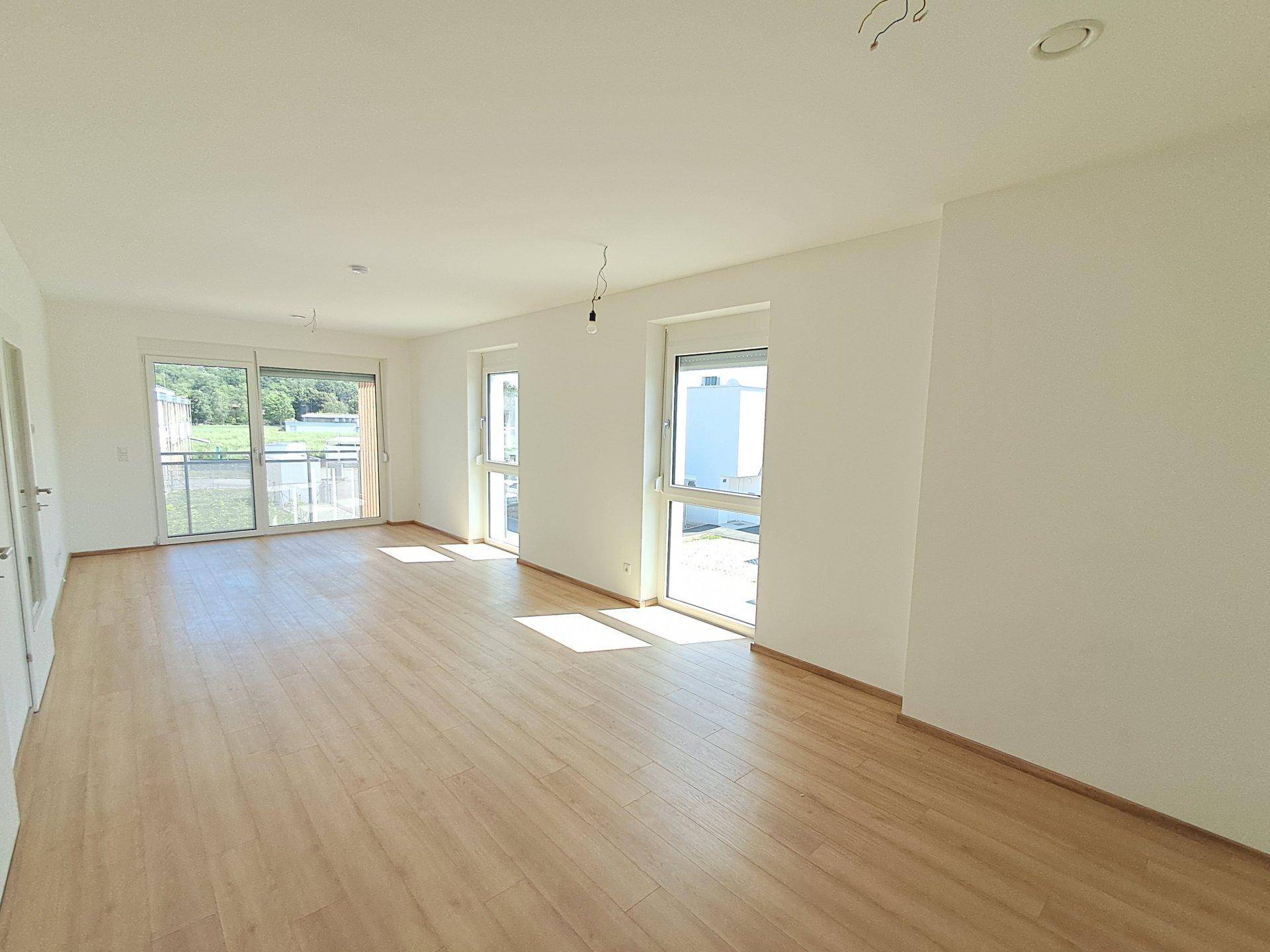 Geräumiger, unmöblierter Wohnbereich mit großen Fenstern und hellem Laminatboden, ideal für viel Tageslicht.