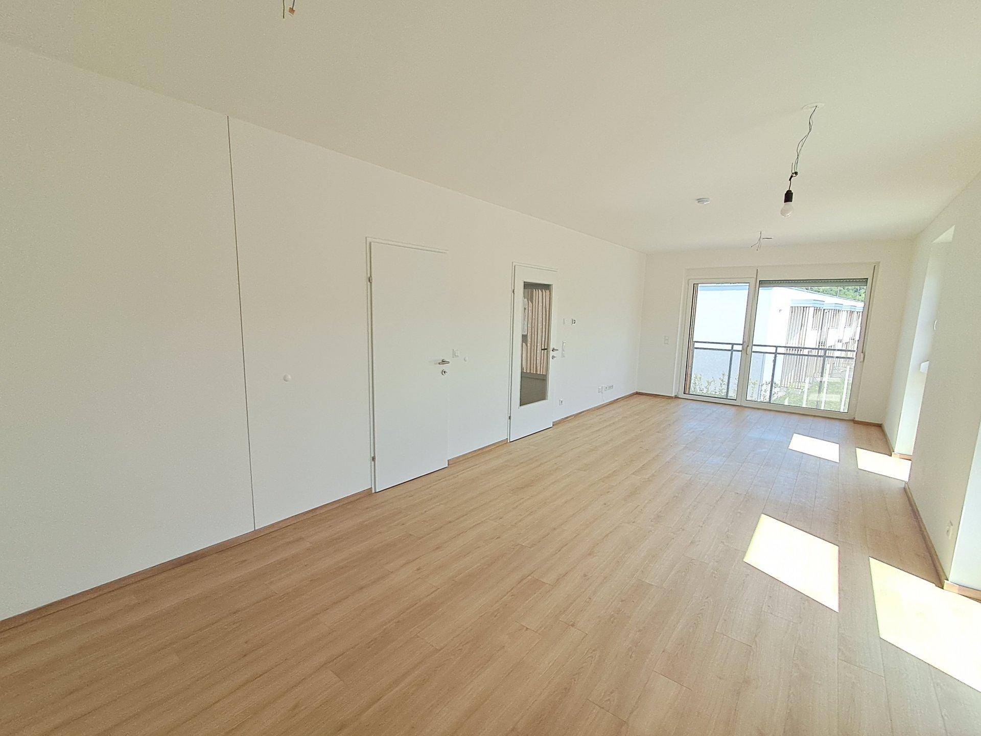 Heller, leerer Wohnbereich mit Laminatboden und Zugang zu einem Balkon, bereit zur individuellen Gestaltung.