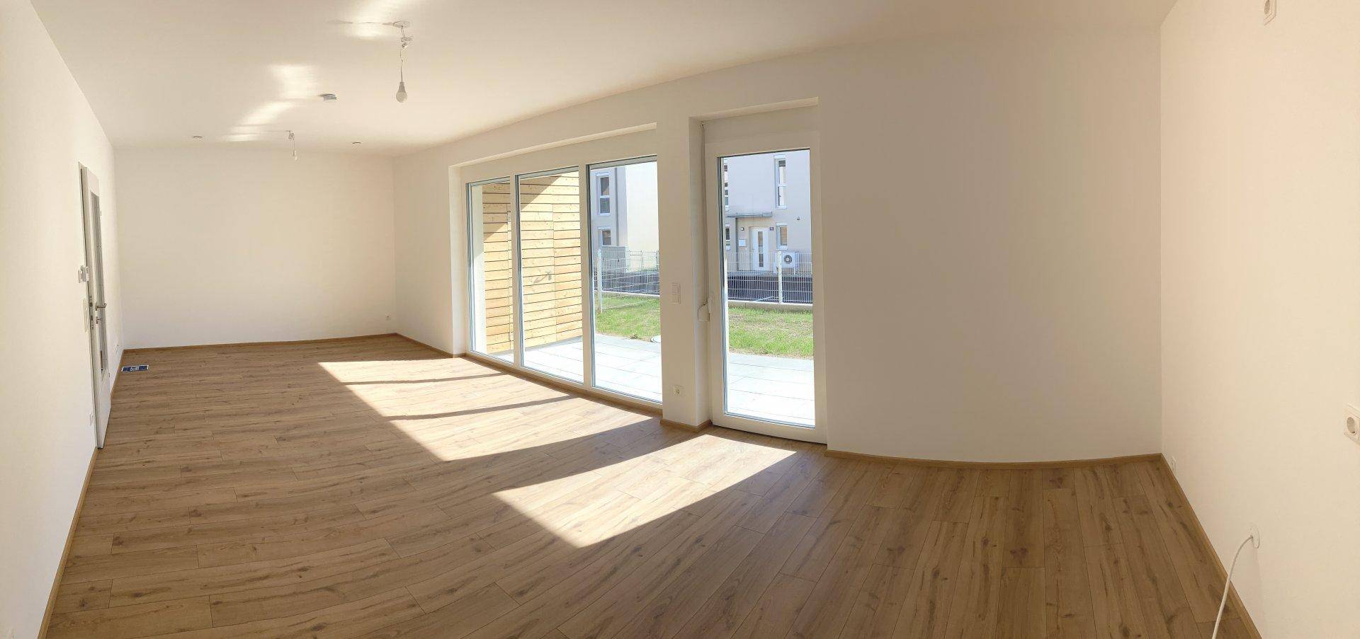 Geräumiger, unmöblierter Wohnbereich mit großen Fenstern und Holzboden, der viel Tageslicht hereinlässt.