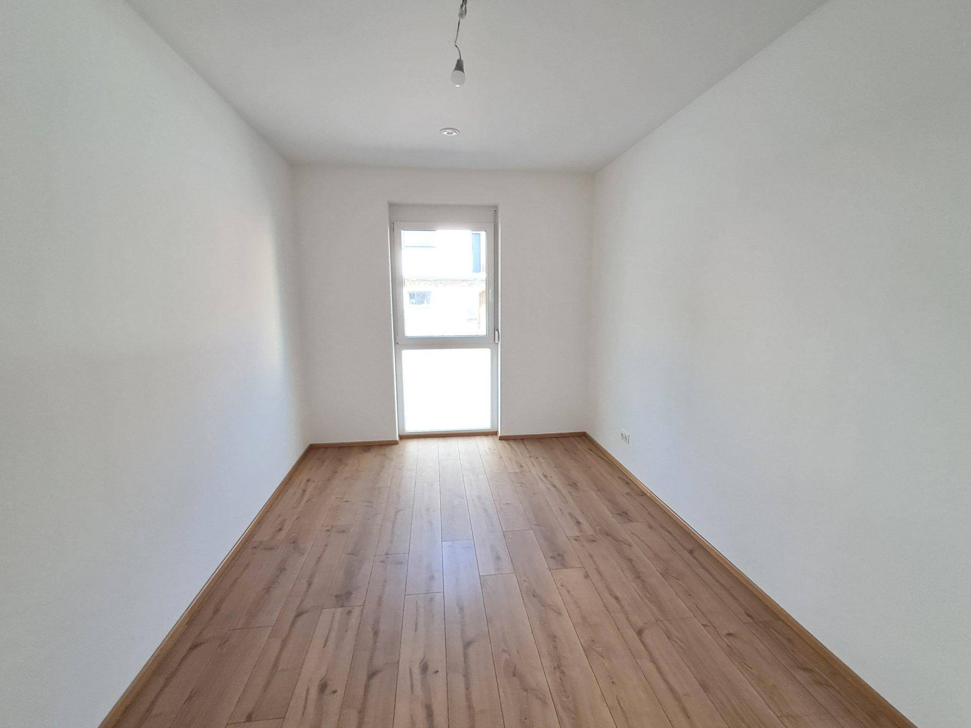 Unmöbliertes Schlafzimmer mit Holzboden und einem Fenster, das viel Tageslicht hereinlässt.