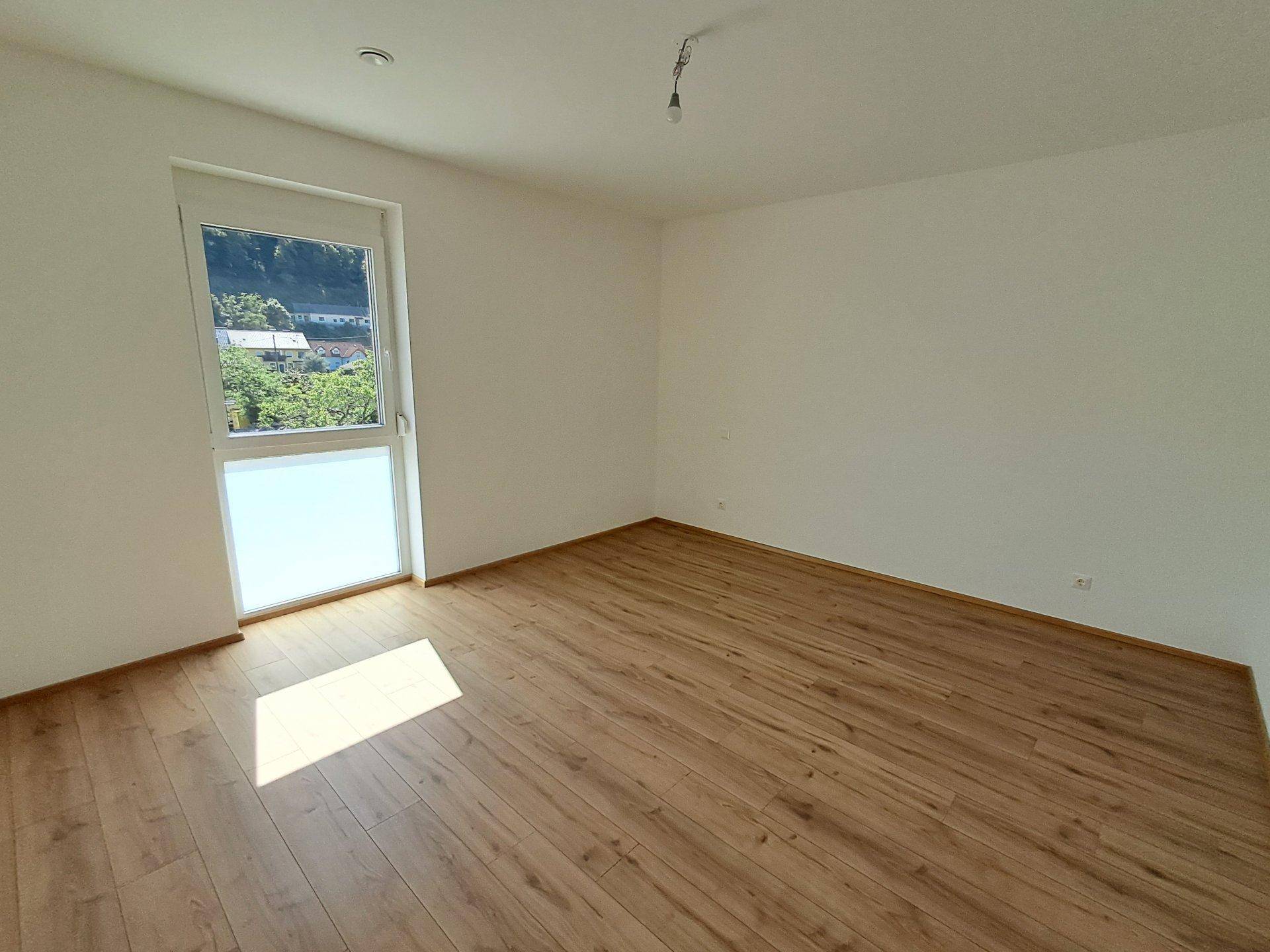 Leeres Schlafzimmer mit Holzboden und einem großen Fenster, das viel Licht hereinlässt.