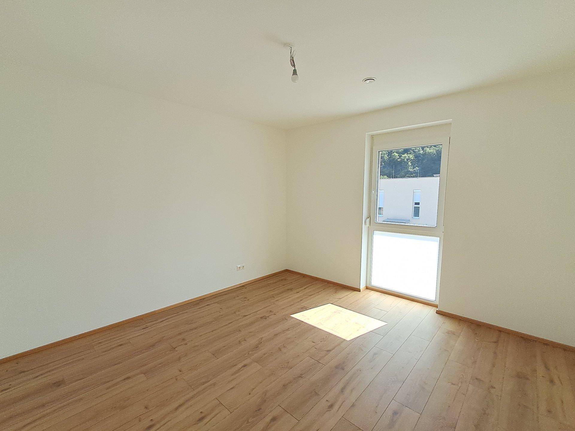 Unmöbliertes Zimmer mit Holzboden und Fenster, das viel Tageslicht hereinlässt.