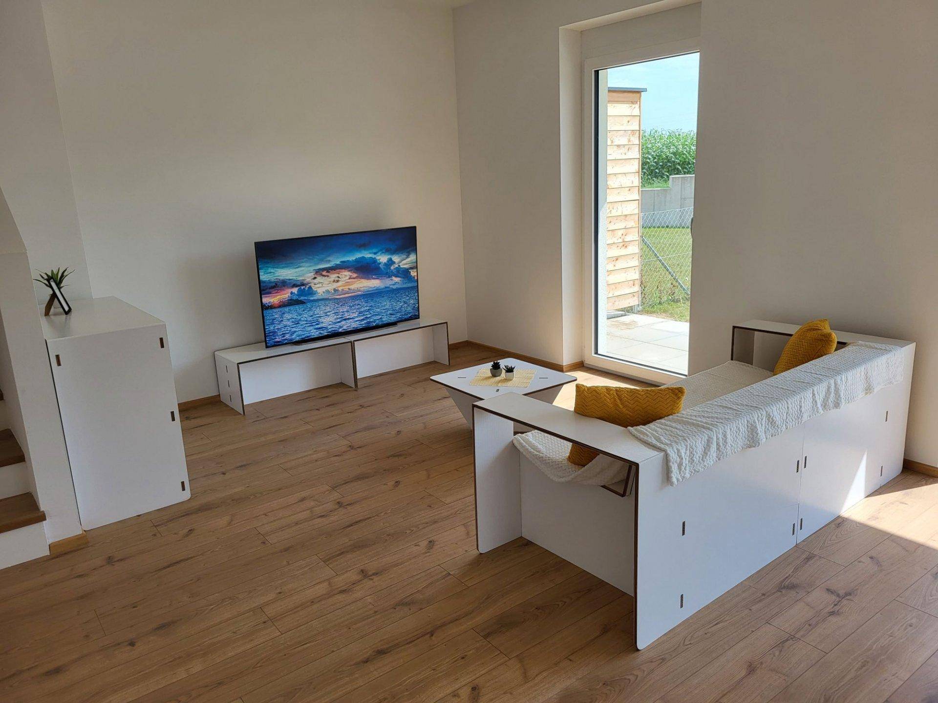 Gemütliches Wohnzimmer mit Laminatboden, Fernseher und Zugang zum Außenbereich durch eine große Tür.
