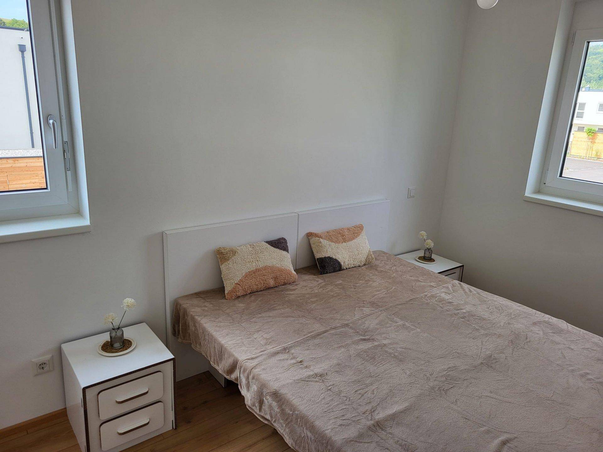Helles Schlafzimmer mit Laminatboden, einem Doppelbett und zwei Fenstern für natürliche Belichtung.