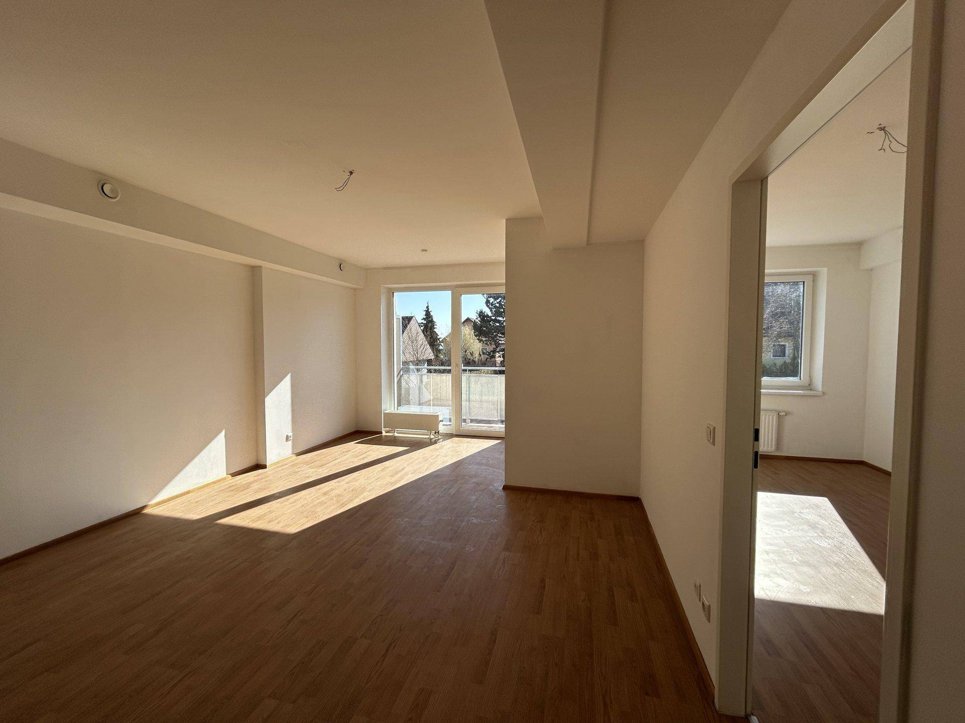 Helles, leeres Wohnzimmer mit Holzboden und Zugang zu einem kleinen Balkon, durchflutet von Sonnenlicht.