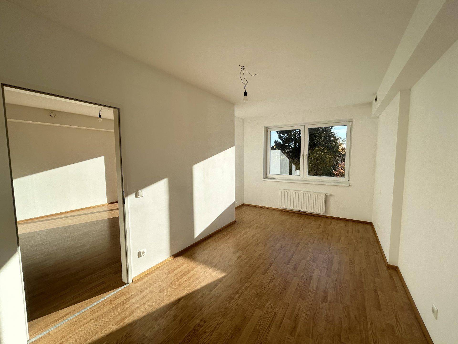 Helles, leeres Zimmer mit Holzboden, weißen Wänden und einem Fenster, das viel Sonnenlicht hereinlässt.