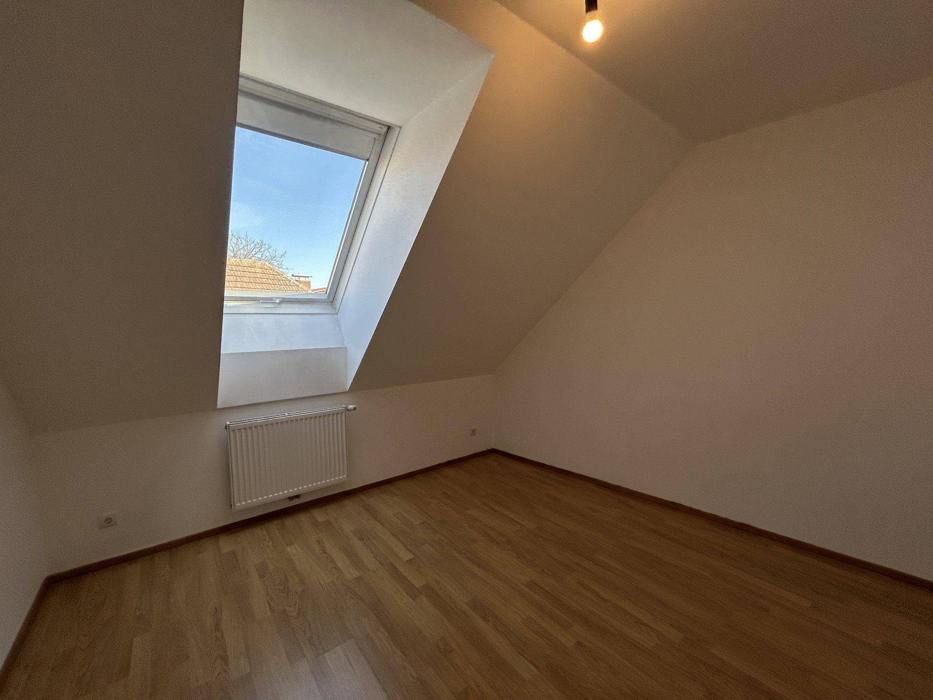 Helles, leeres Zimmer im Dachgeschoss mit Holzboden, Dachfenster und Heizkörper.