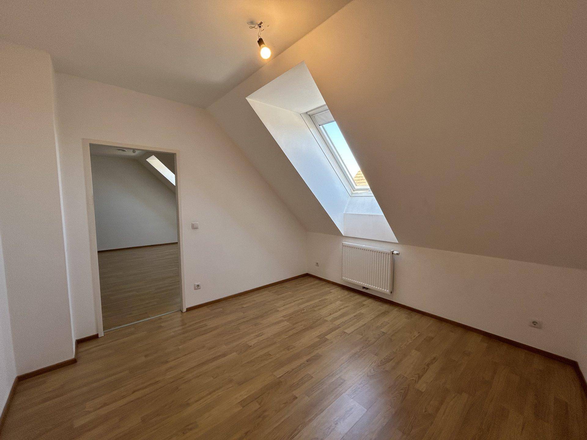 Leeres Zimmer im Dachgeschoss mit Holzboden, Dachfenster und Zugang zu einem weiteren Raum.