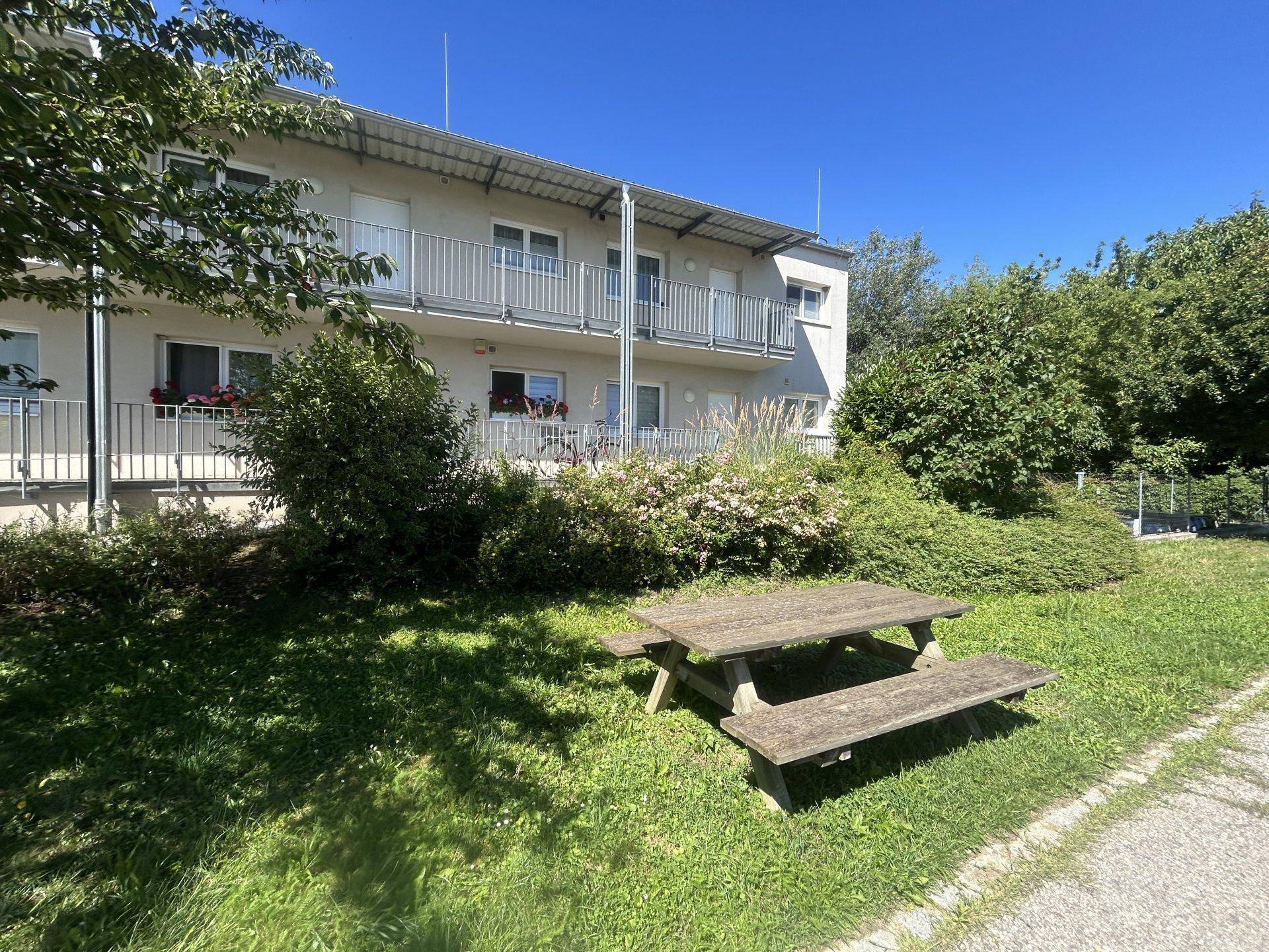 Rückseite eines Wohnhauses mit mehreren Balkonen und einem Gemeinschaftsgarten mit Picknicktisch im Grünen.
