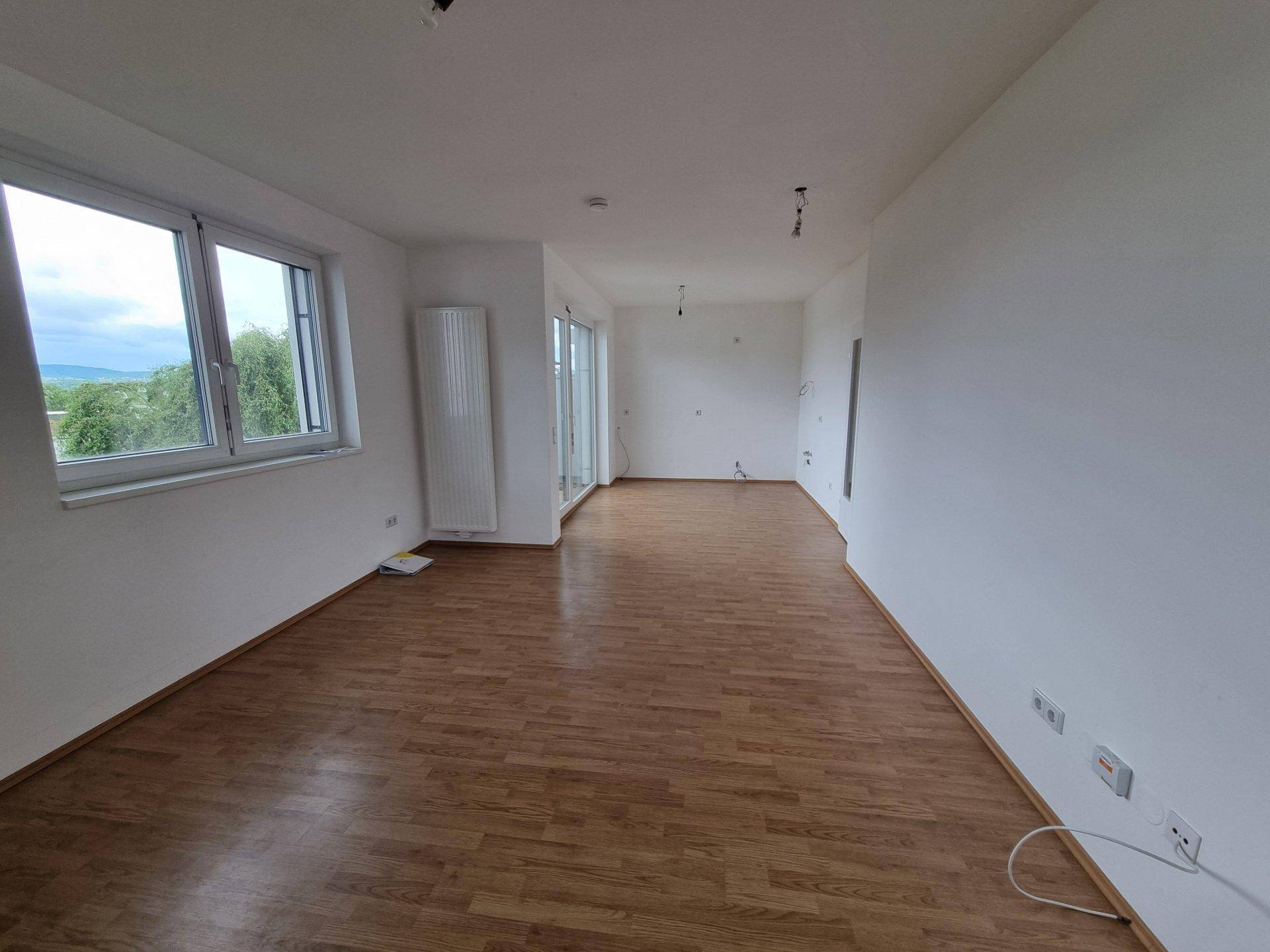 Helles, leeres Wohnzimmer mit Laminatboden, großen Fenstern und Zugang zu einem Balkon.