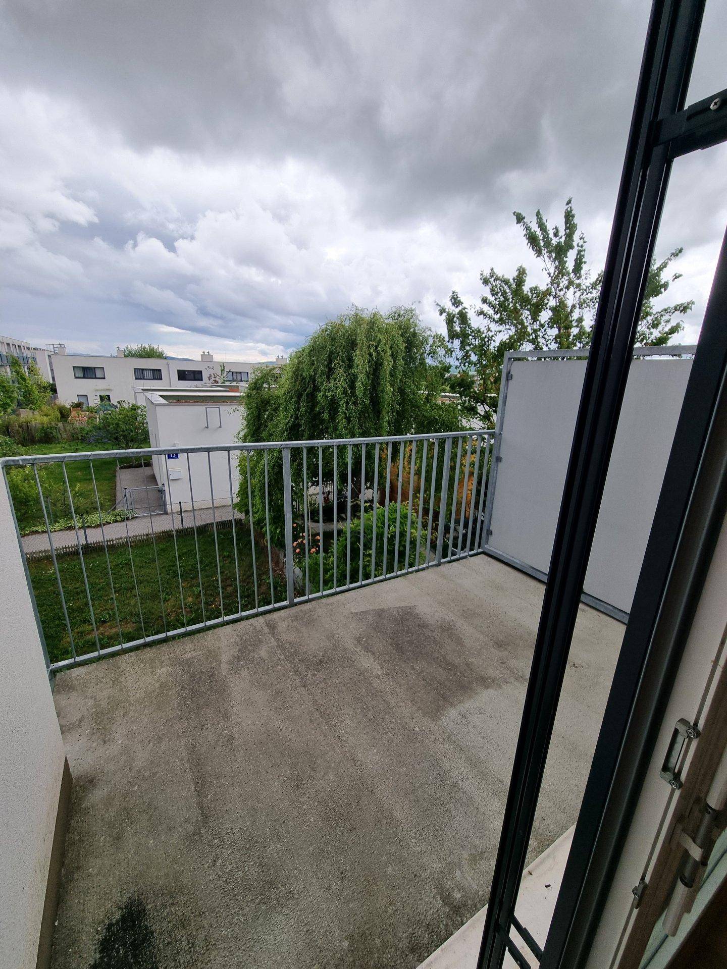 Balkon mit Betonboden und Metallgeländer, bietet Blick auf Grünflächen und umliegende Gebäude.