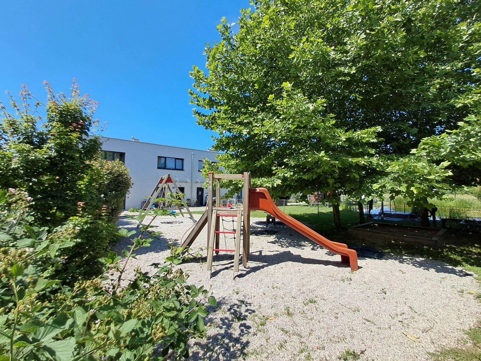 Kinderspielplatz mit Rutsche und Schaukeln, umgeben von Bäumen und Grünflächen der Wohnanlage.