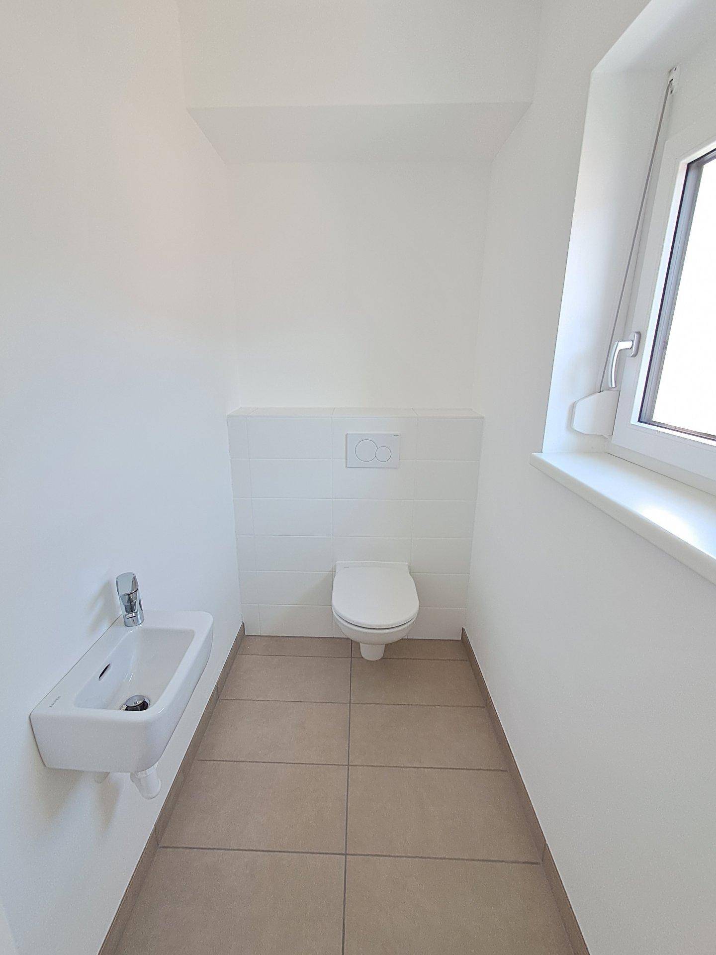 Helles Gäste-WC mit wandhängendem WC, kleinem Waschbecken und Fenster für natürliche Belüftung.