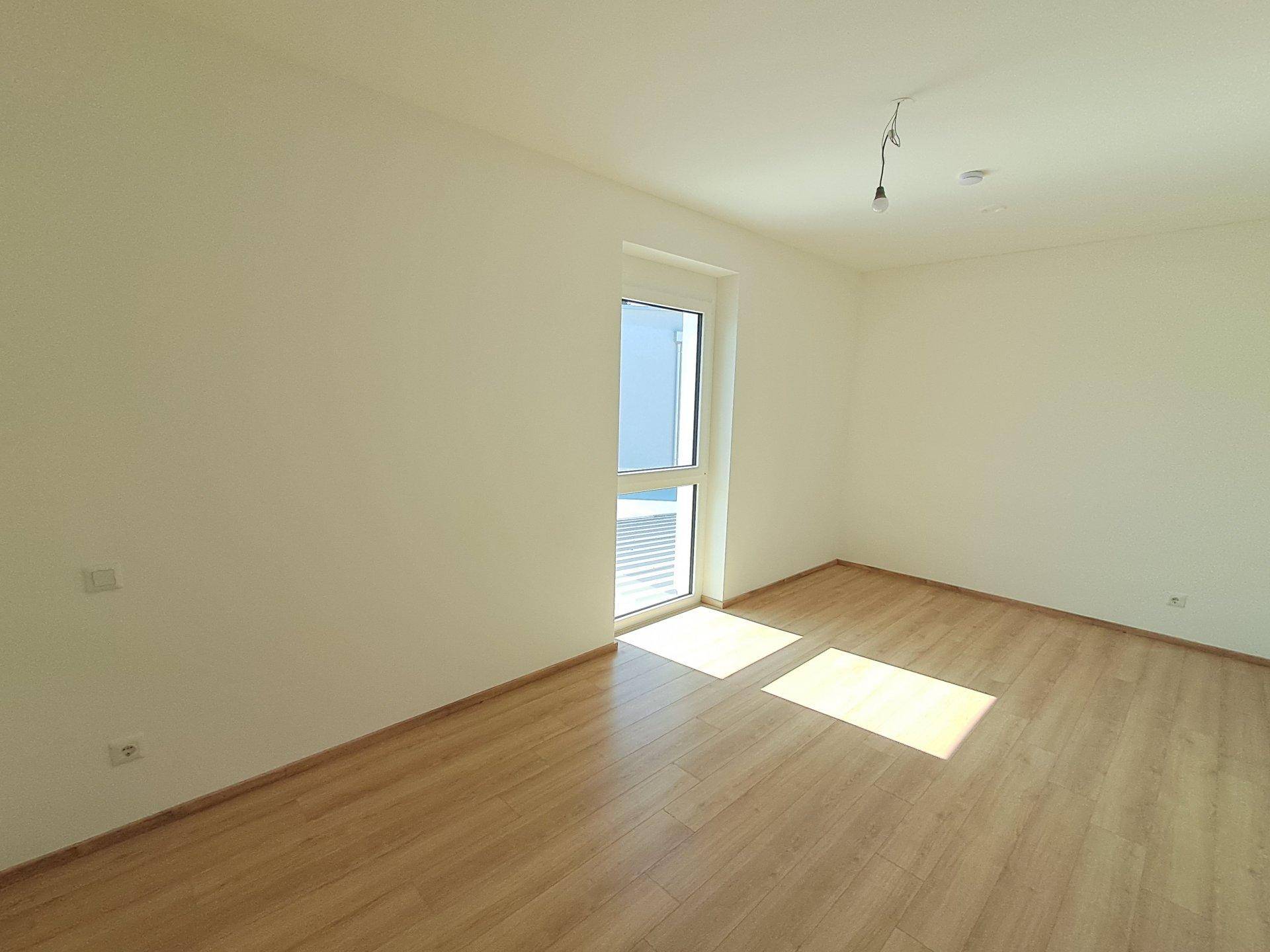Helles, leeres Schlafzimmer mit Laminatboden und großem Fenster, bereit zur individuellen Gestaltung.