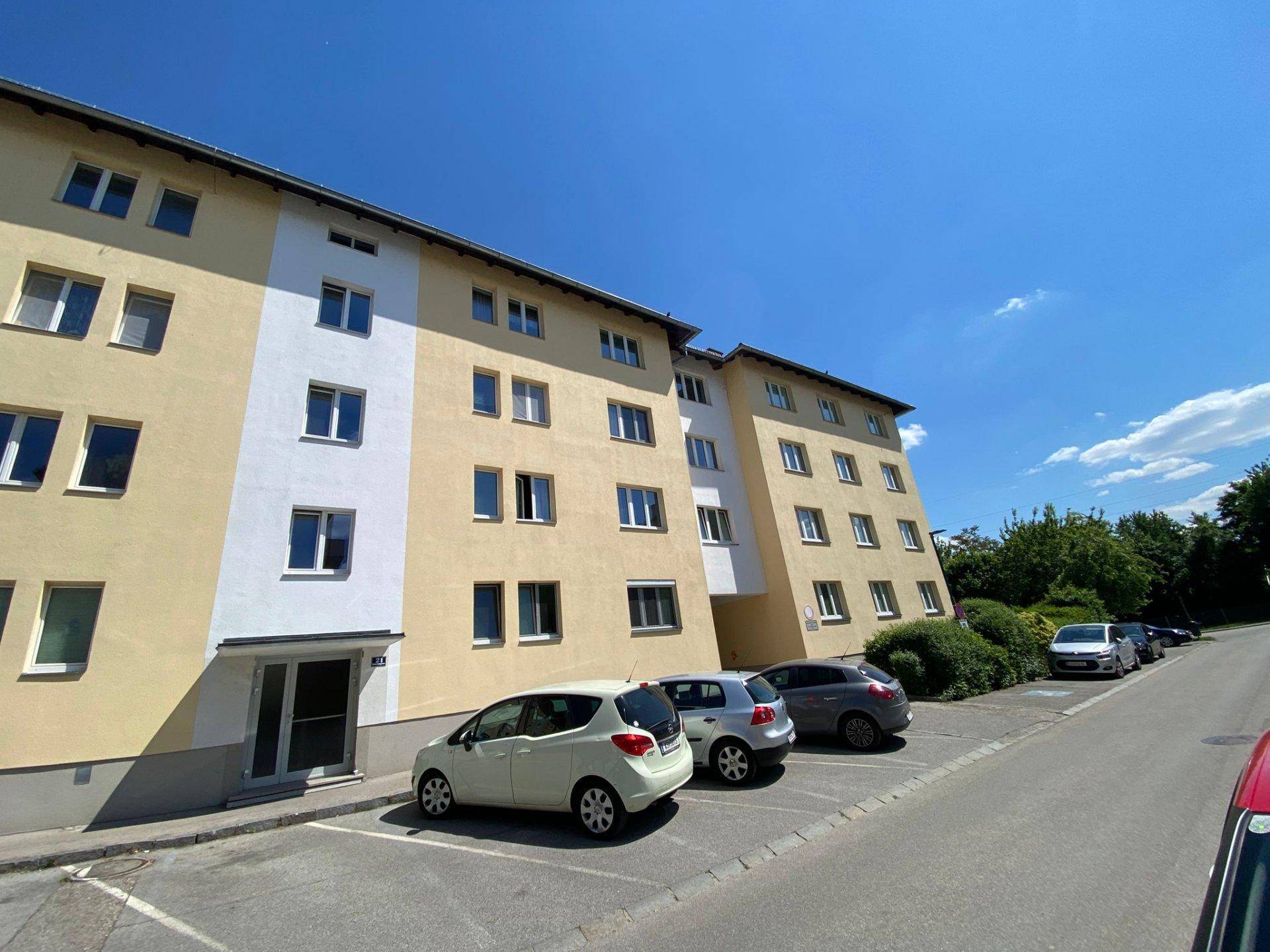 Mehrstöckiges Wohngebäude mit Parkplätzen davor und einer Straße unter blauem Himmel.