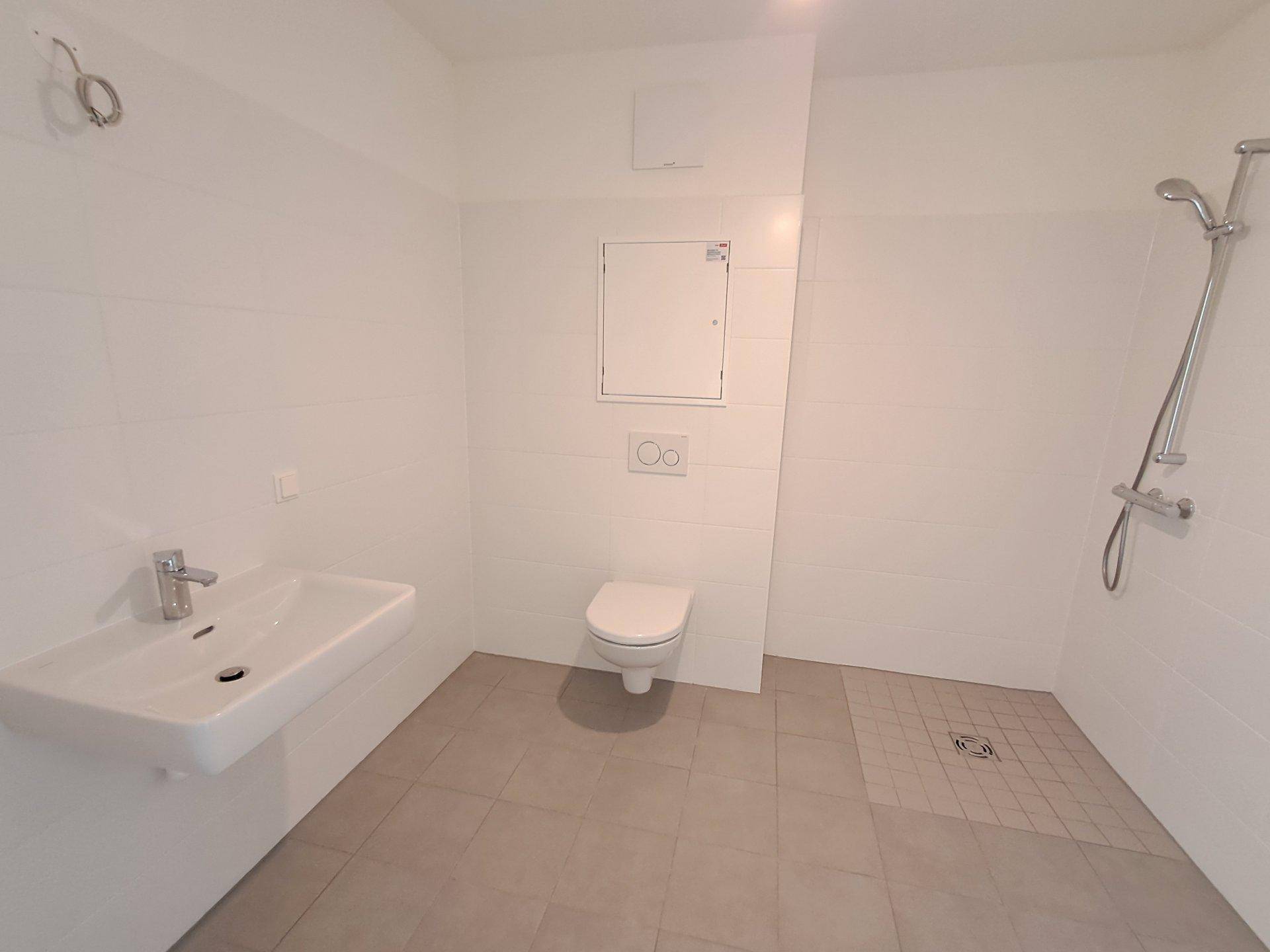 Helles, unmöbliertes Badezimmer mit Waschbecken, WC und Duschbereich.