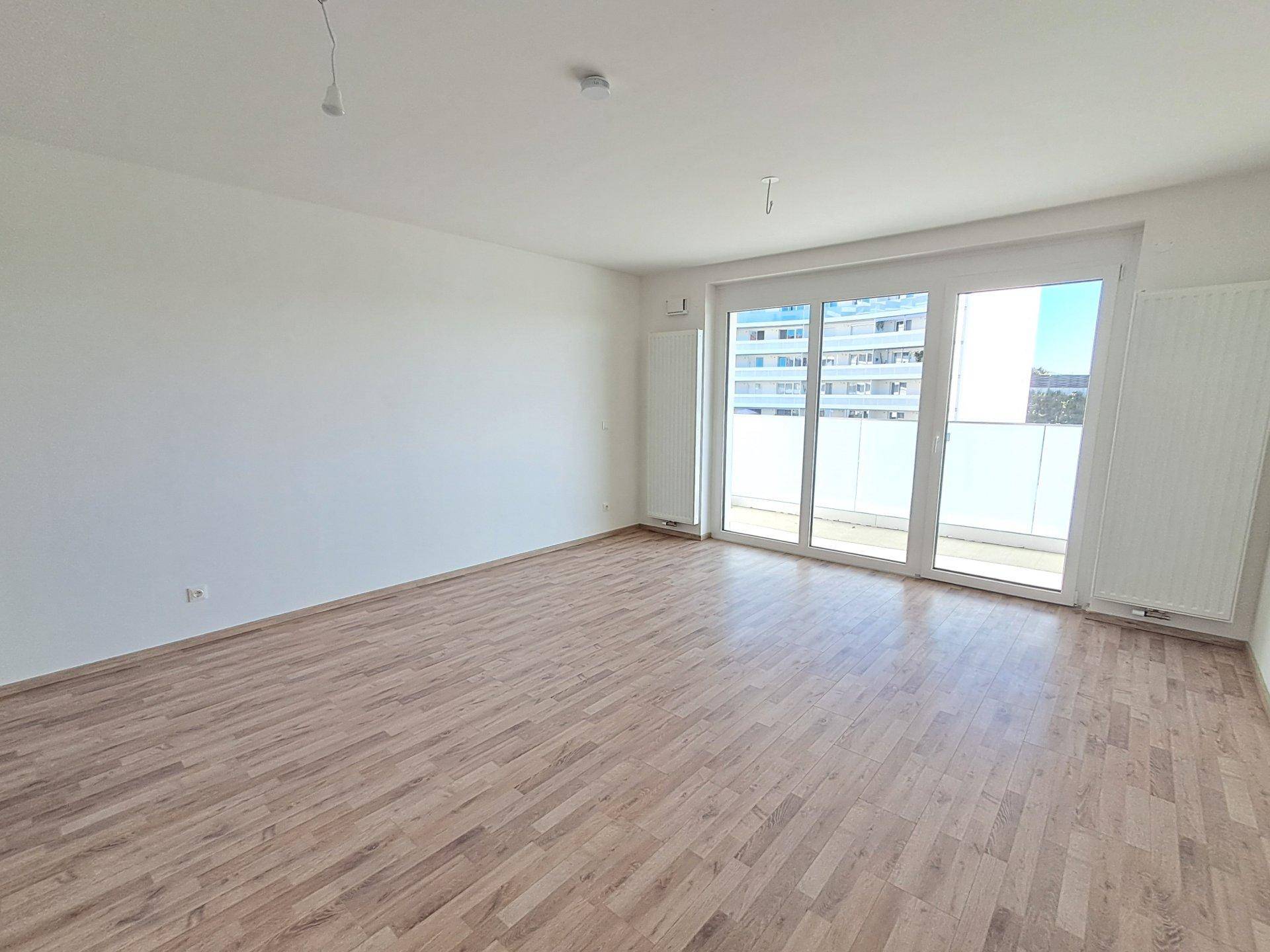 Lichtdurchfluteter Raum mit Holzboden und breiter Balkontür, Blick auf umliegende Gebäude.