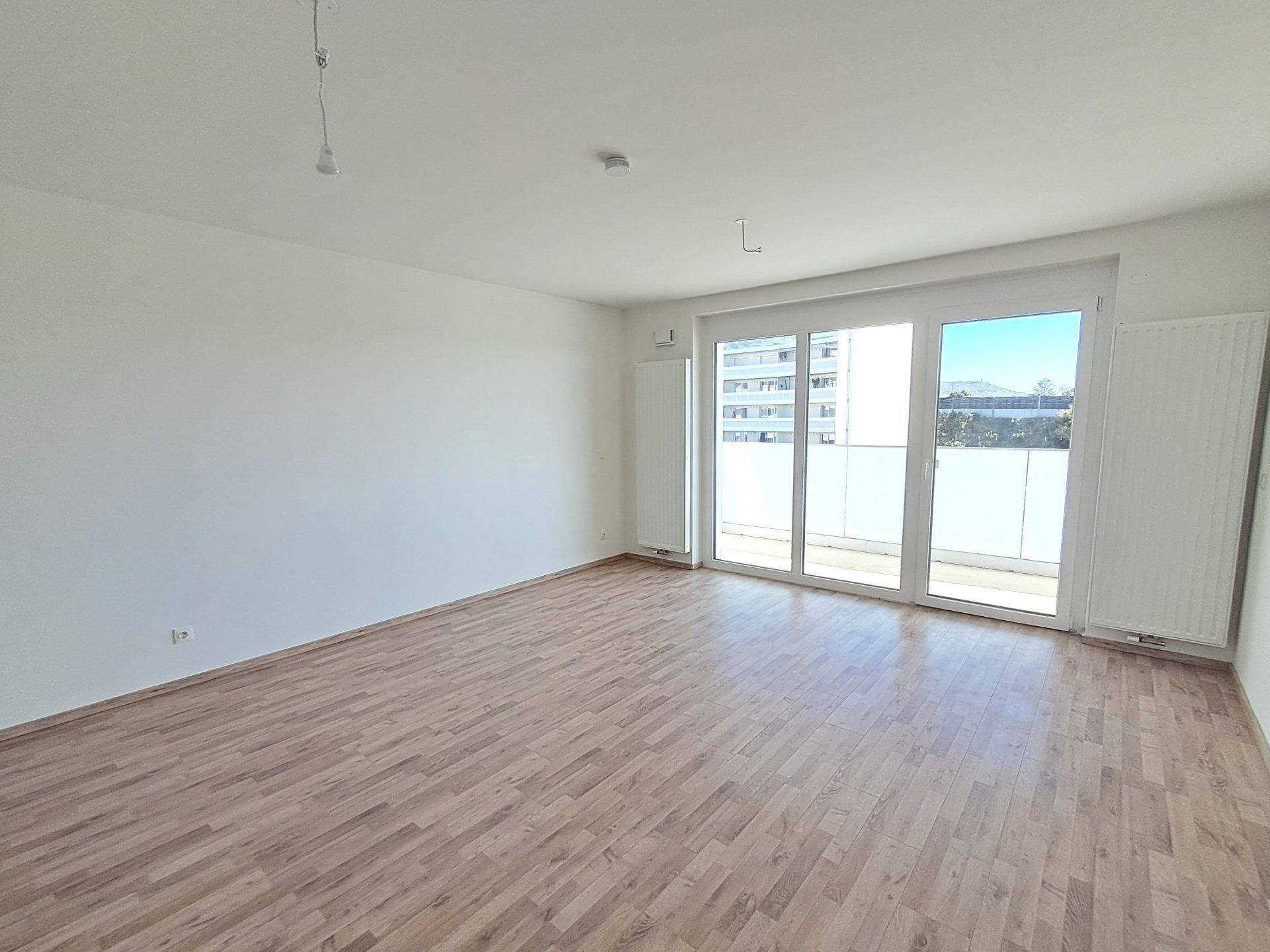 Leerer Wohnbereich mit Holzboden, großen Fenstern und Blick auf die Umgebung.