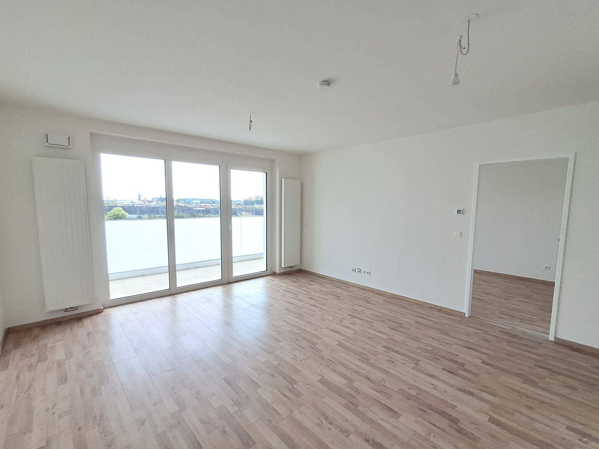 Heller, leerer Raum mit Holzboden und großen Fenstern, die Zugang zu einem Balkon bieten und den Raum erhellen.