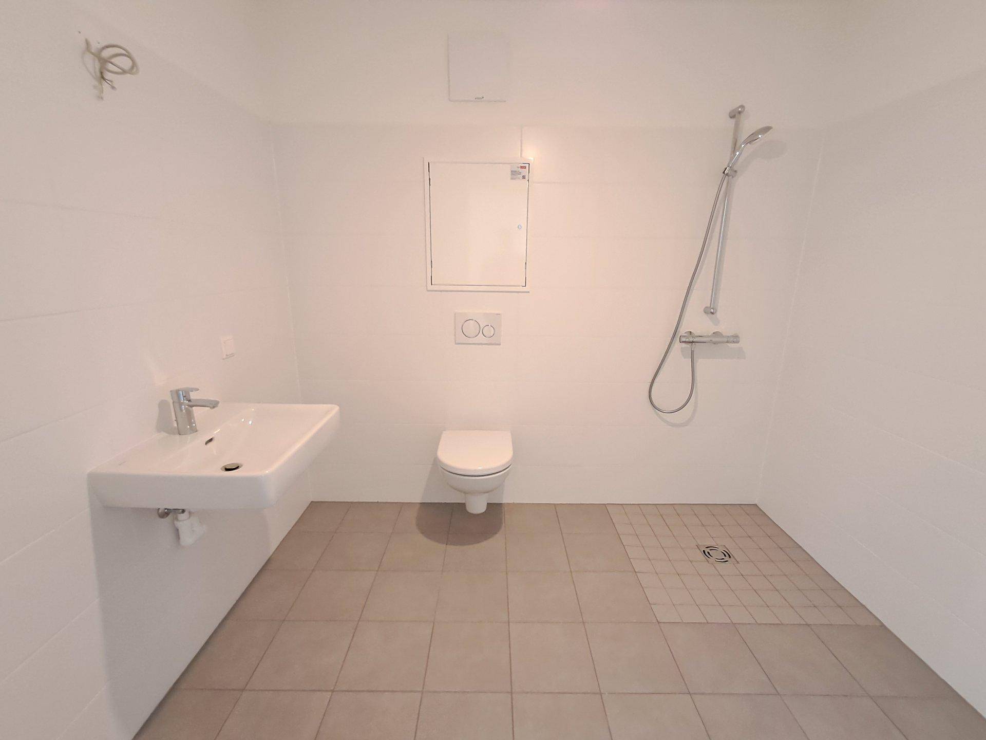 Helles, unmöbliertes Badezimmer mit wandhängendem WC, Waschbecken und einer geräumigen Duschfläche.